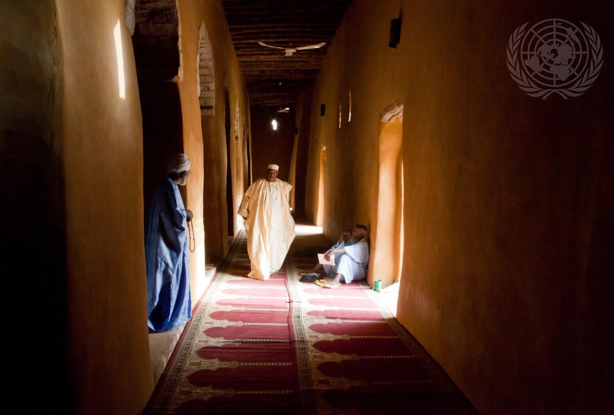 Drie mannen in een huis. Links staat een man in een blauw gewaad. In het midden loopt een man in een wit gewaad door de gang. Rechts zit een man op de grond.