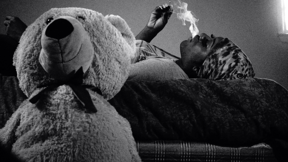 Een van de hoofdpersonen uit de documentaire ligt op bed. Ze rookt een sigaret. Naast haar staat een teddybeer.