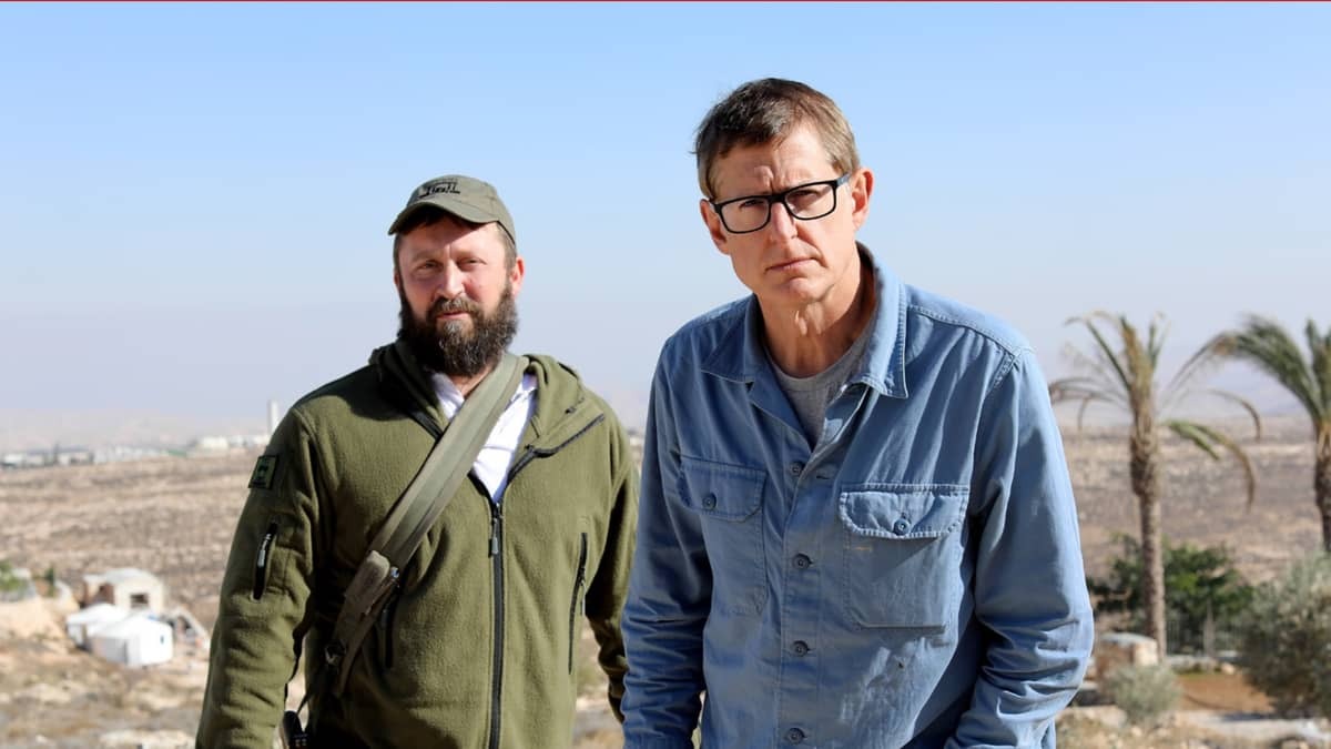 Louis Theroux en een man in een groen vest staat in een warme omgeving, palmbomen op de achtergrond.