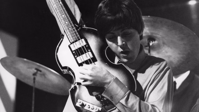 Een archiefbeeld van Paul McCartney met zijn basgitaar. Het is een zwart-wit foto en hij is heel jong. Hij heeft donker haar en zijn bas heel hoog naast zijn hoofd en hij speelt erop.