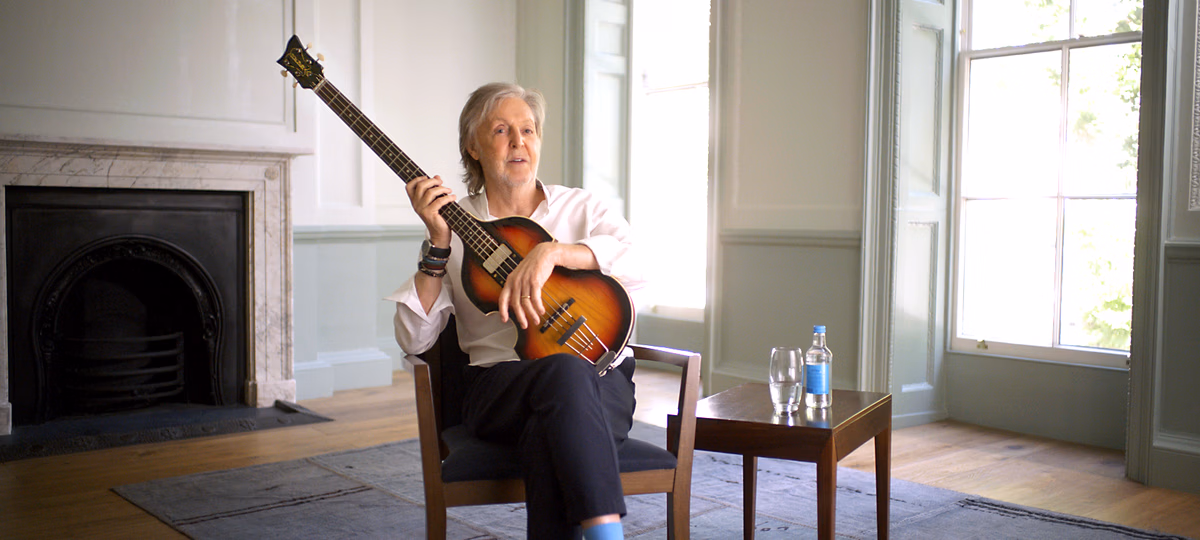Paul McCartney met zijn bas. Hij zit op een stoel in een grote lege ruimte en kijkt naar de camera. Het is duidelijk een interviewsetting. Hij heeft zijn basgitaar in zijn handen.