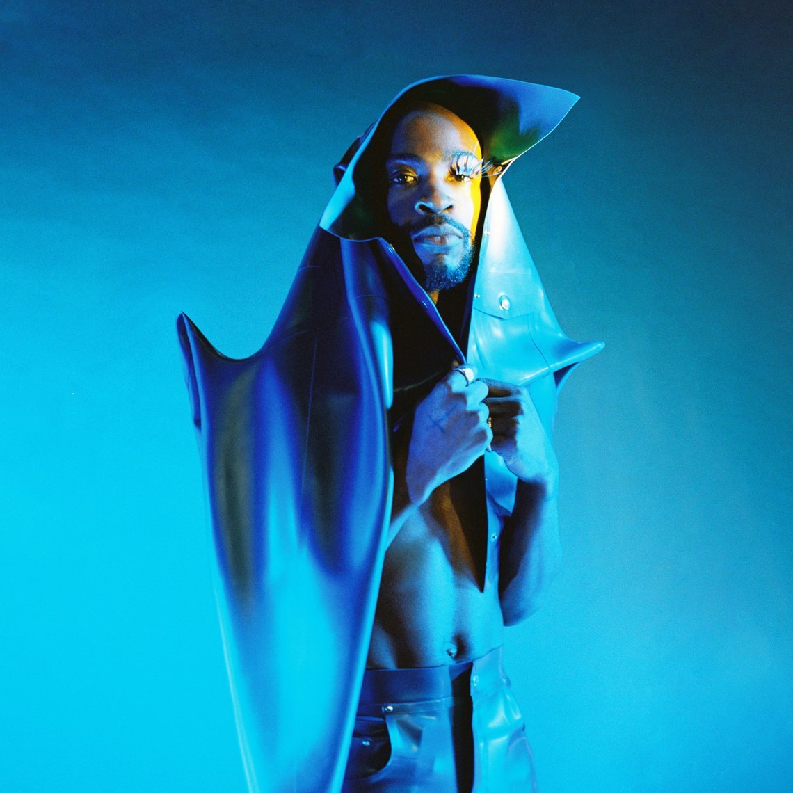 Artiest Isaï staat in blauw licht voor een blauwe achtergrond. Om zich heen draagt hij een soort sculptuur-achtige jas. Hij kijkt recht in de camera, bij zijn rechteroog draagt hij grote nepwimpers.