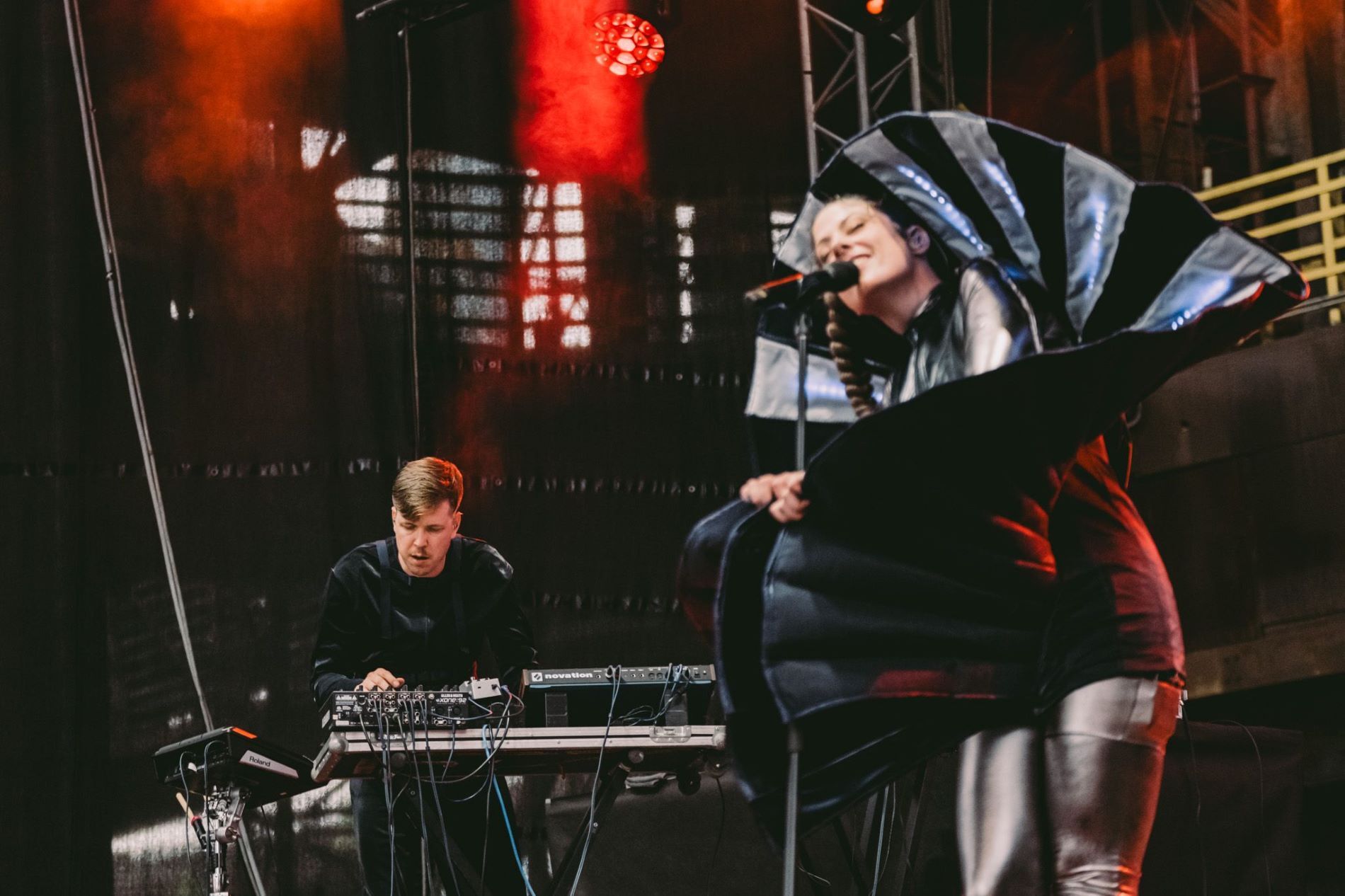 Foto van twee mensen die aan het optreden zijn. Samen vormen ze een band genaamd CUT_. Links draait een witte man achter een paneel met knoppen. Hij is in focus, heeft bruin haar en kijkt geconcentreerd. Rechts zingt een vrouw met haar ogen dicht. Ze is niet on focus en draagt een alienachtige outfit die helemaal om haar heen is gewikkeld. Het lijkt alsof ze uit een soort blad komt.