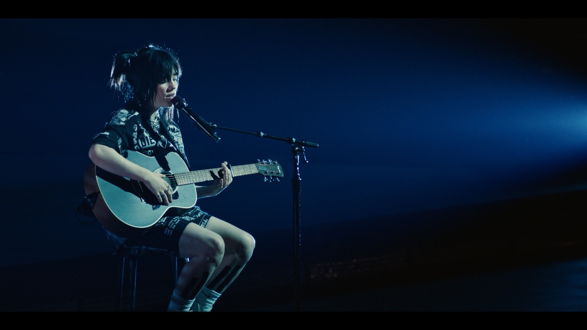Billie eilish op een krukje met een akoestische gitaar in haar handen, voor een microfoon. De spotlight is op haar gericht en het geeft een blauwig licht.