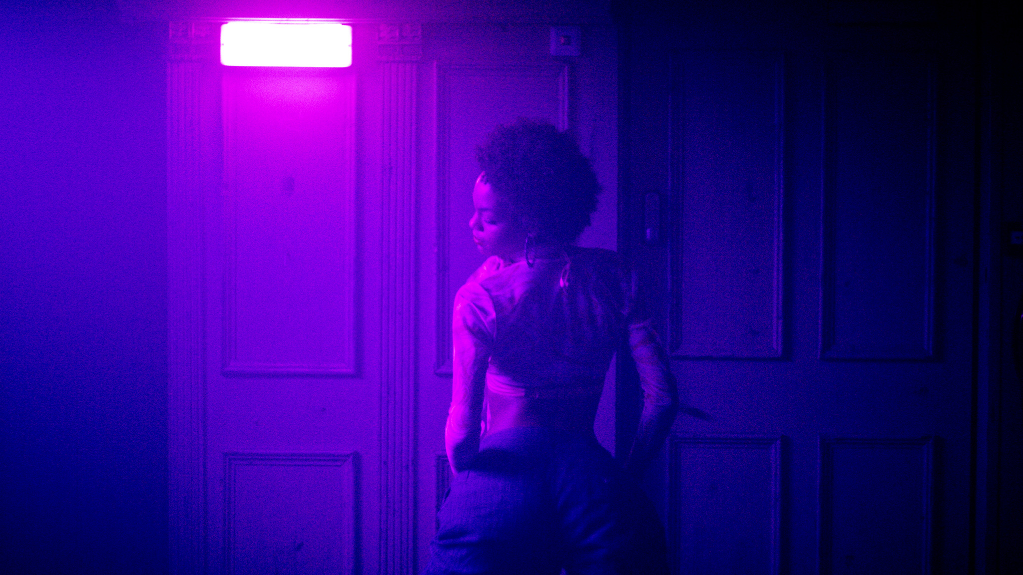 Vrouw met kort krullend haar danst op bubbling-muziek. Ze staat voor een witte deur en de ruimte is blauw en paars verlicht. De vrouw staat met haar rug naar de camera gericht, maar we zien wel de zijkant van haar gezicht.
