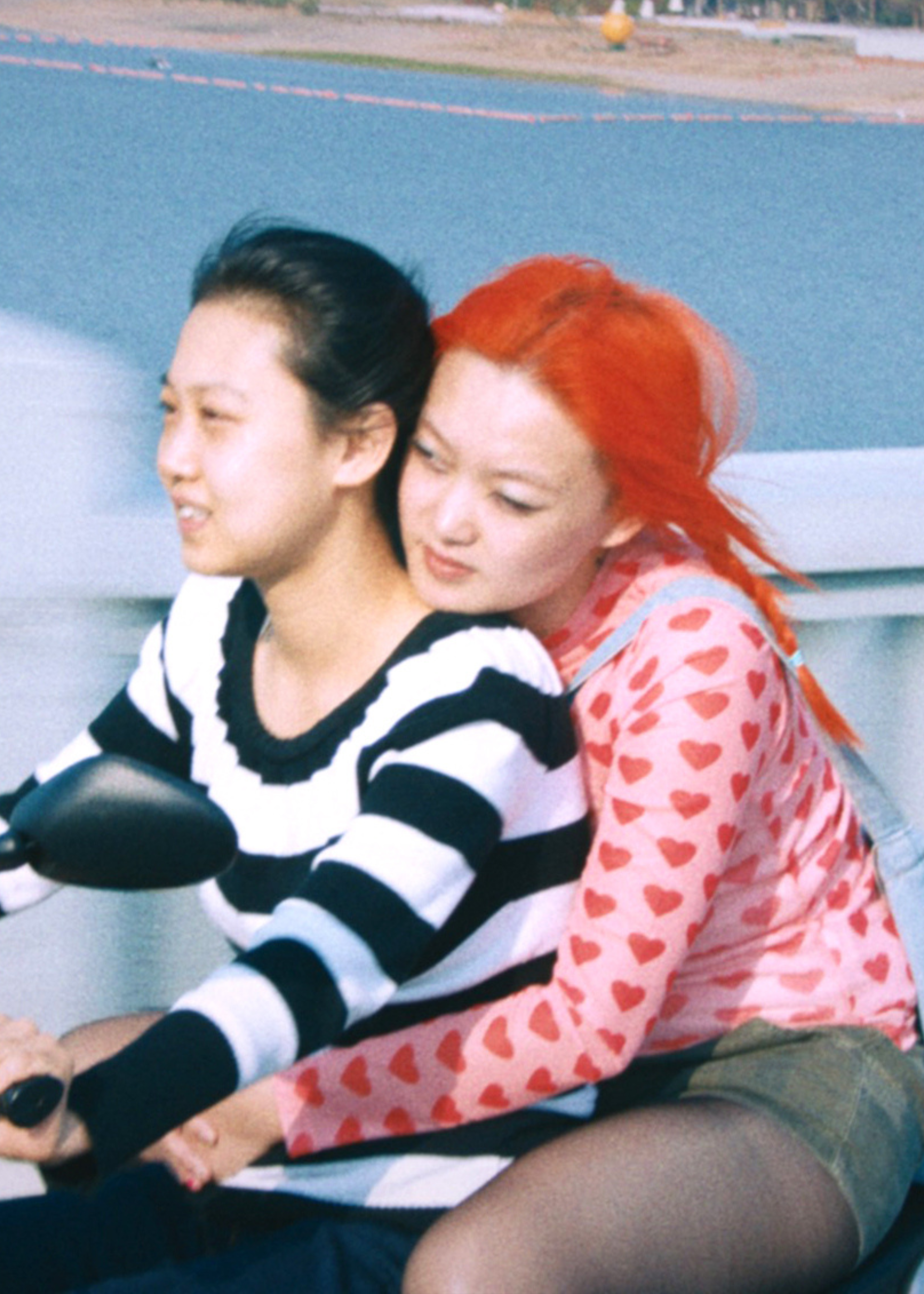 Twee Chinese actrices uit de film 1 Girl Infinite zitten samen op een scooter. Degene die rijdt, draagt een zwart/wit gestreept shirt en heeft zwart haar. Degene die achterop zit, houdt de andere persoon vast om haar middel. Ze draagt een roze shirt met rode hartjes, een groen rokje en heeft felrood haar.
