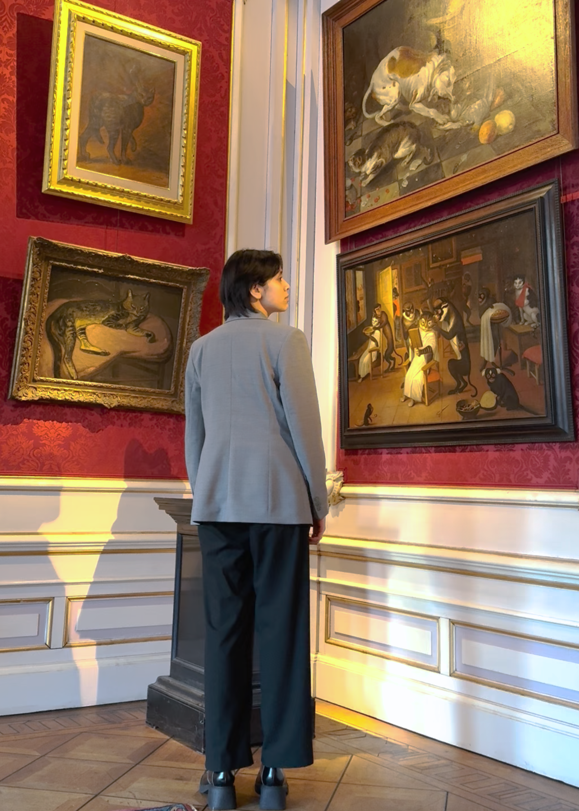 Redacteur Seshiekela staat voor een hoge muur in een Amsterdams grachtenpand. Aan de muur hangen allerlei oude schilderijen met kattenkunst eraan.