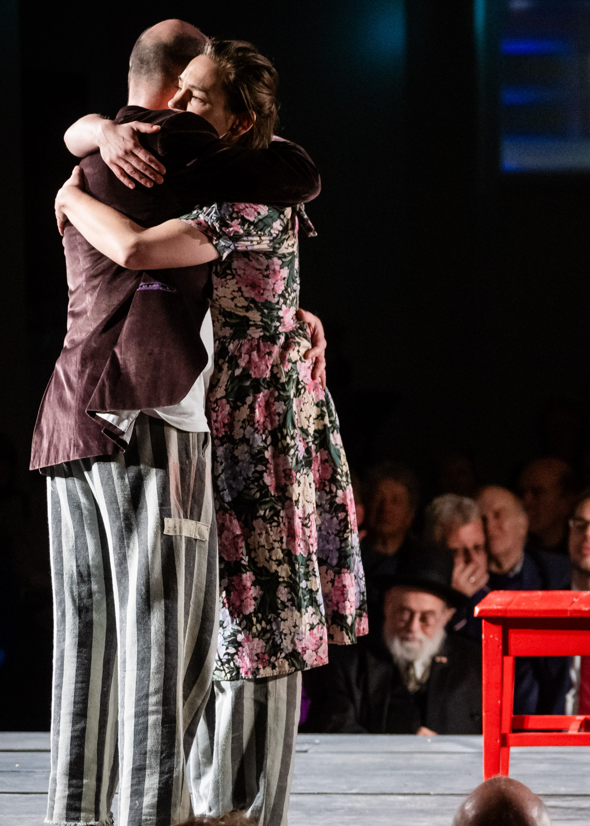 Op de foto zijn twee knuffelende personen op een podium te zien. Een persoon, de vrouw, heeft een bloemetjesjurk aan tot over haar knieën. Daaronder draait ze een loszittende grijs-gestreepte broek. De man draagt een bruin colbert. Ook hij draait dezelfde soort grijs-gestreepte broek. Naast hen op het podium staan een rode tafel en stoel.