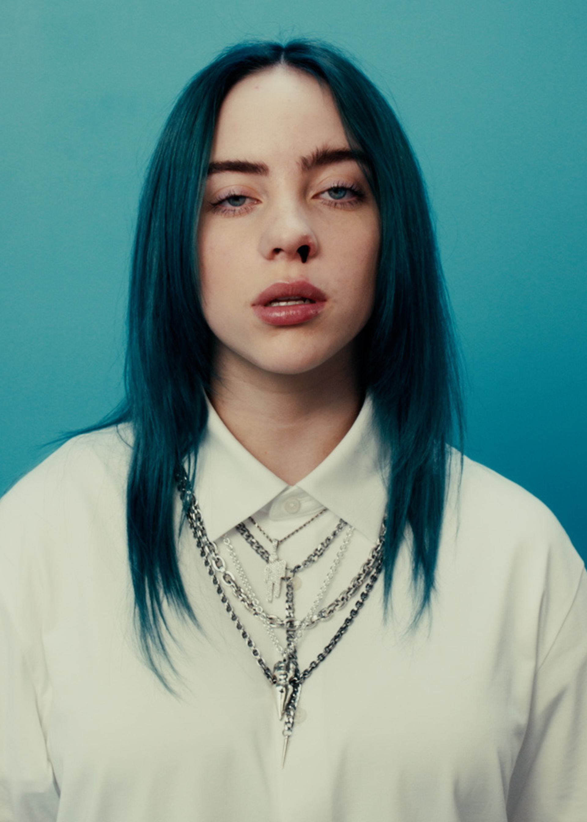 Zangeres Billie Eilish staat voor een blauwe muur. Ze heeft blauw haar, draagt een wit overhemd met kettingen erover en er komt bloed uit haar neus.