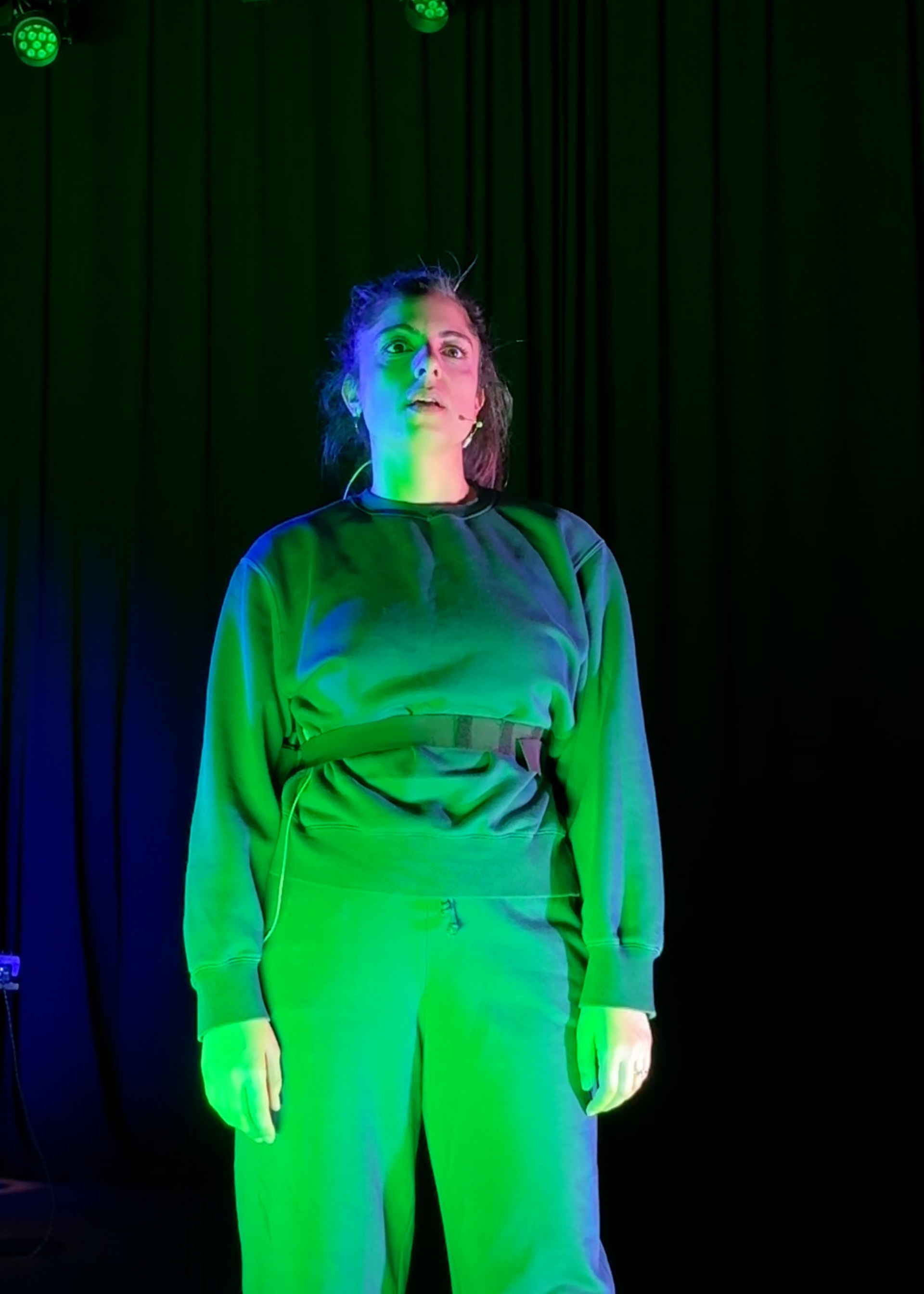 Een actie foto van Yeliz op het toneel. Ze heeft een blauw pak aan en staat in fel groen licht. Ze kijkt serieus en vlak langs de camera.
