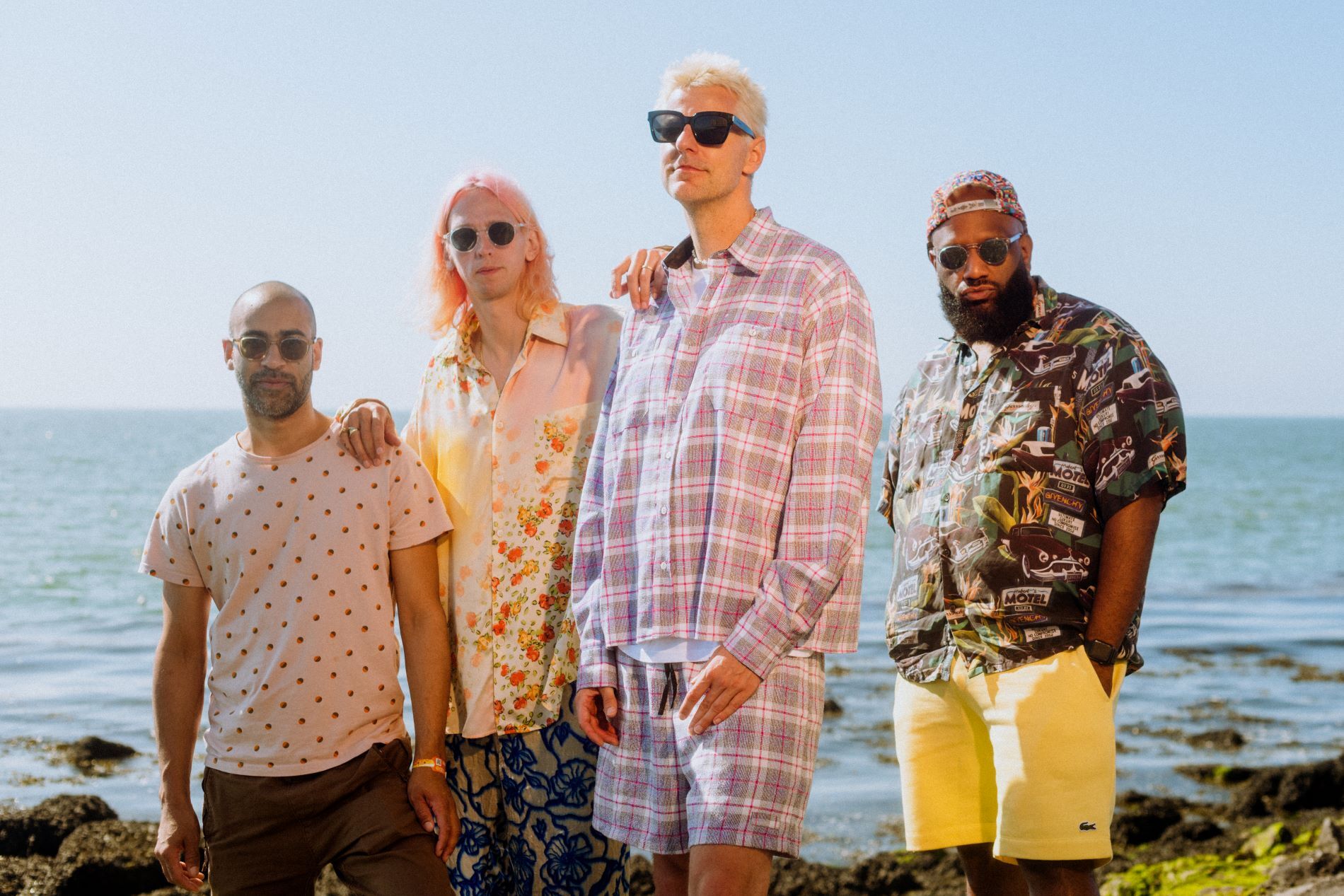 Kleurenfoto van de band De Jeugd van Tegenwoordig. Vier mannen: links een met een gestippeld shirt en een zonnebril, daarnaast een man met roze haar en een bloemetjesbroek en-shirt. In het midden (rechts) een man met een zonnebril en een geruit matching pak. Helemaal rechts een man met een pet, zonnebril en overhemd met auto's.