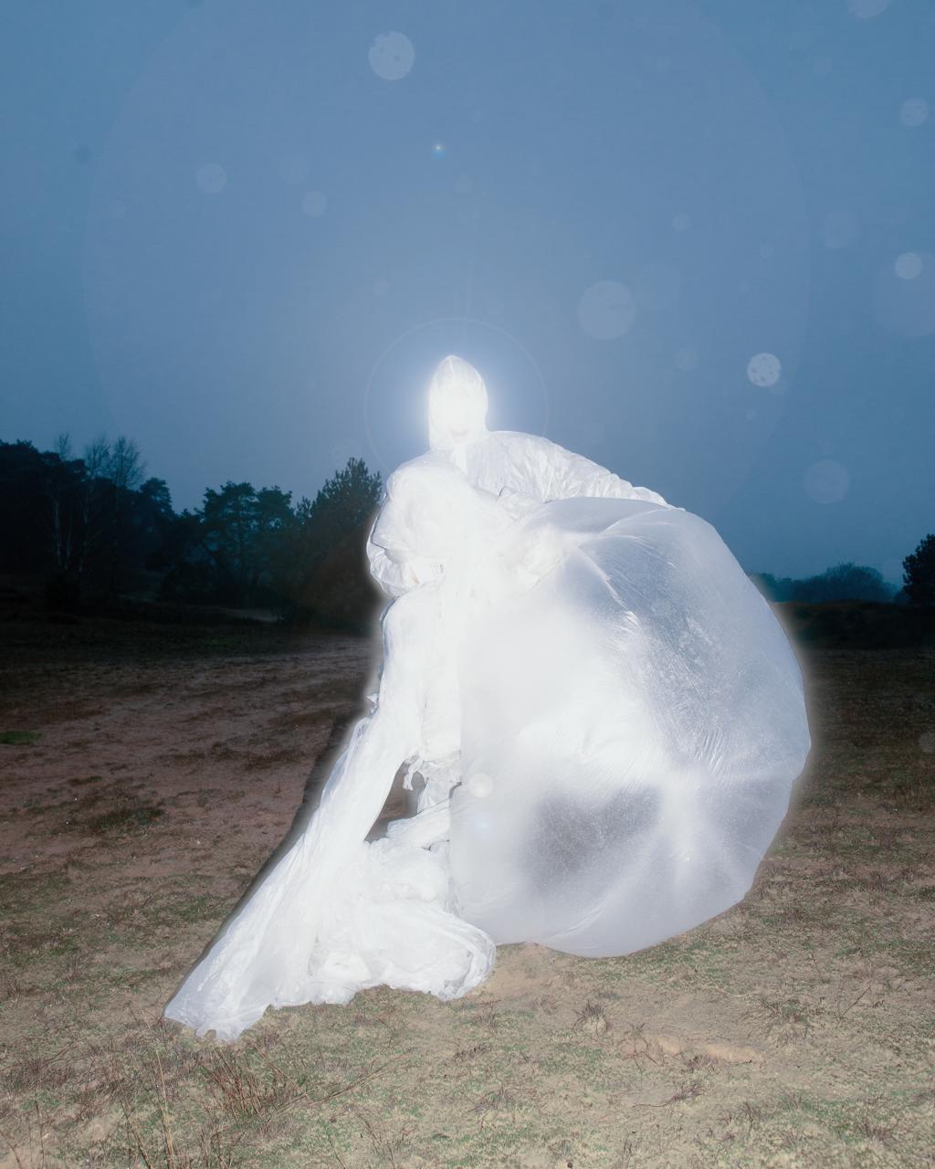 Een mens gehuld in een wit gewaad van materiaal dat op plastic lijkt. Je ziet niets van het gezicht. Op de achtergrond staan bomen.