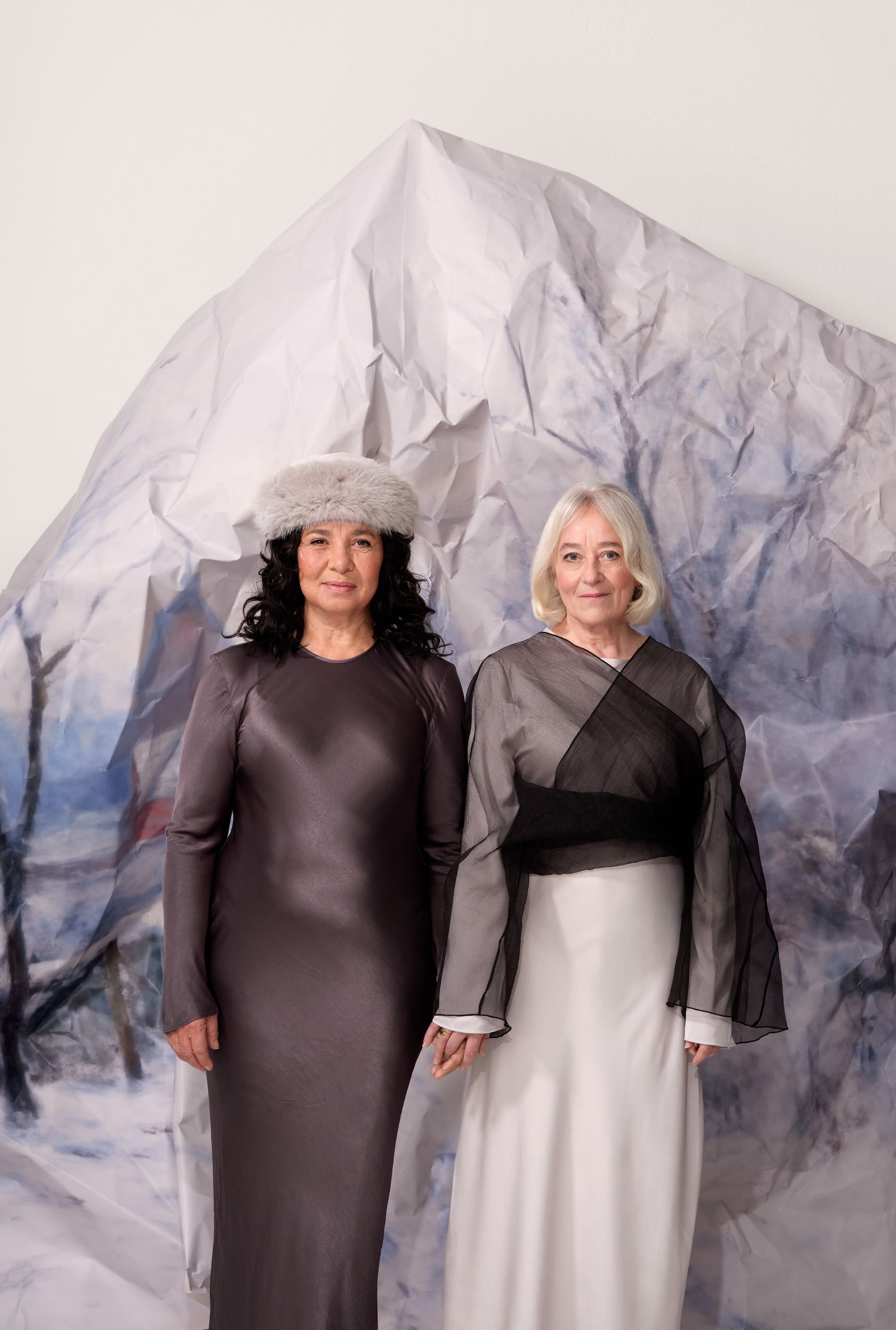 Twee vrouwen staan naast elkaar met hun vingers in elkaar verweven. Achter hen staat een ijsberg van papier. De linker vrouw heeft donkerbruin krullend haar, draagt een grijze muts en een lange bruine jurk. De rechter vrouw heeft kort, stijl en blond haar en draagt een lange zwart/witte jurk.