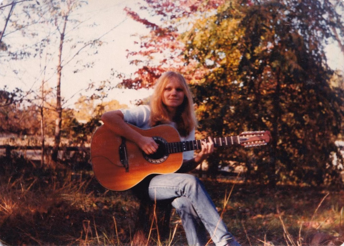 Foto van Eva Cassidy die buiten tussen de bomen zit met een gitaar op schoot. Ze lacht naar de camera.
