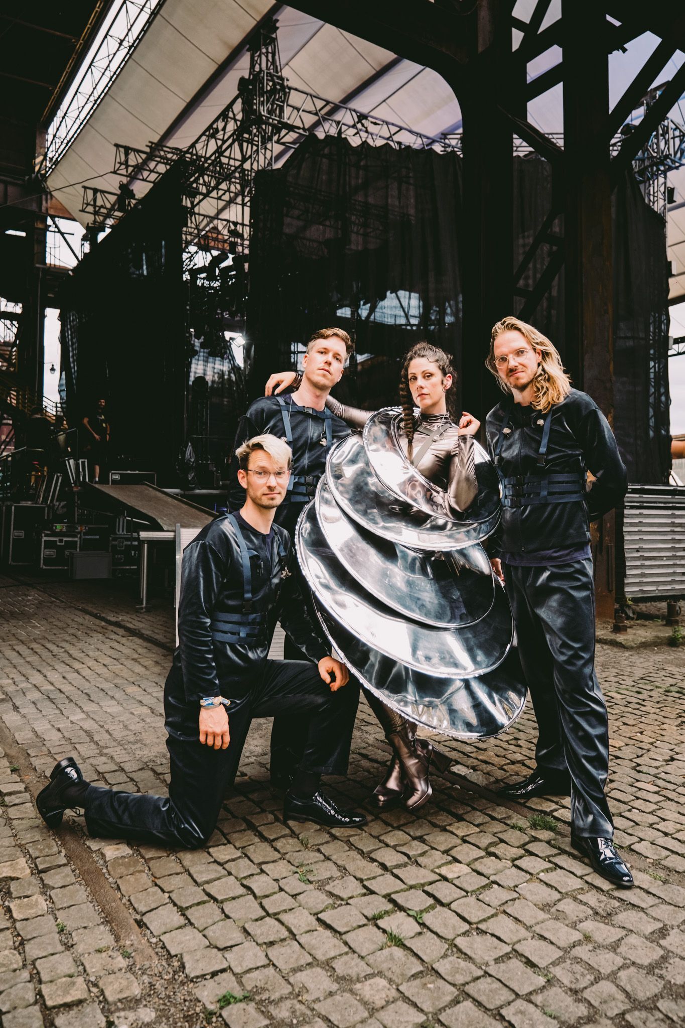 Een foto van vier mensen: links twee witte mannen, in het midden een witte vrouw en rechts ook een witte man. De mannen links hebben beide kort haar, dragen een blauw harnas en een zwart pak. De man rechts draagt dezelfde outfit en heeft lang blond haar. De vrouw in het midden draagt een outfit die lijkt te bestaan uit verschillende zilveren schotels. Haar lange bruine haar draagt ze in een vlecht. Hun blikken zijn serieus.