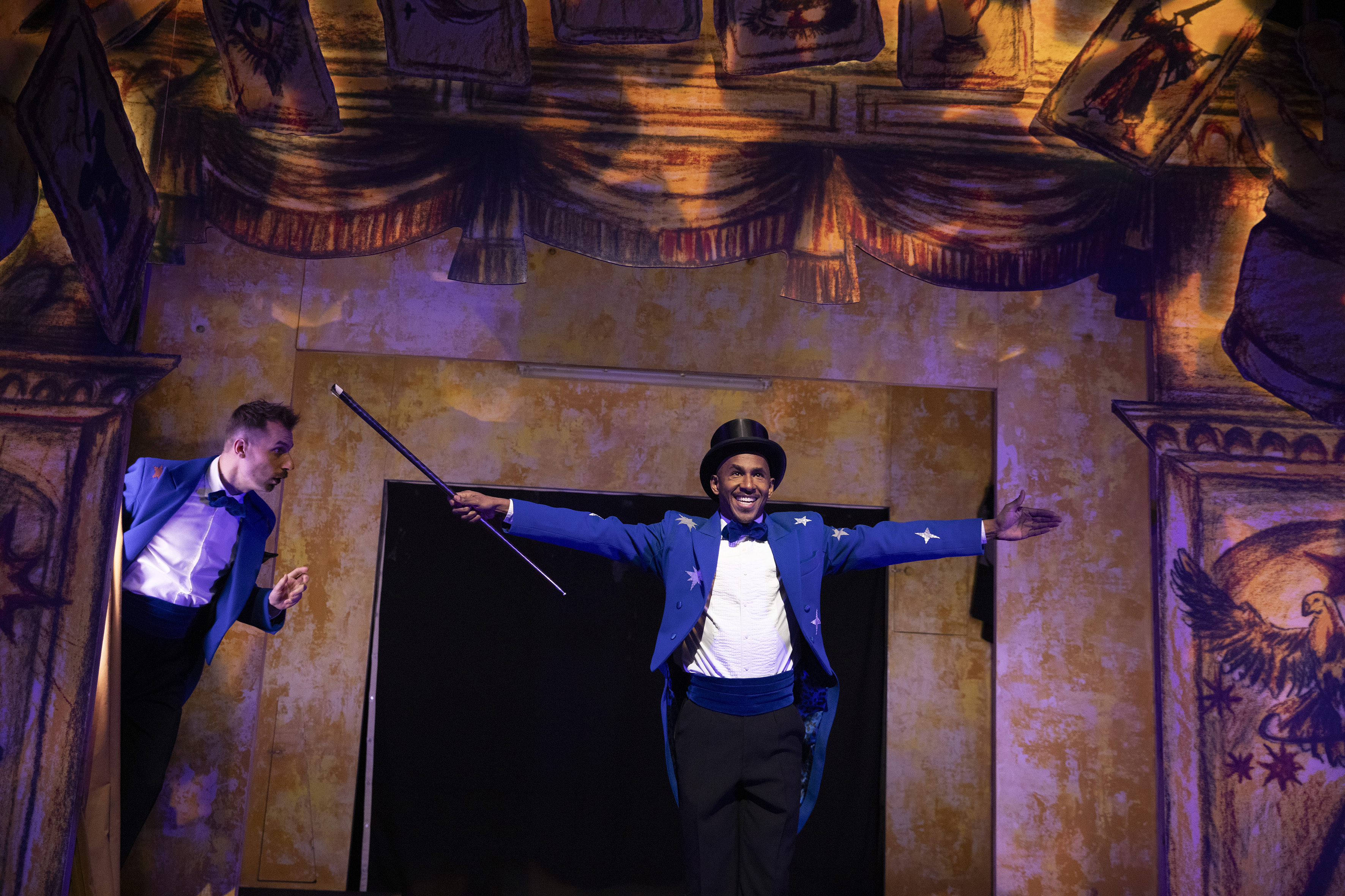 Hier zie je José Montoya met een blauw rokkostuum aan en een broek met een blauwe band om zijn middel. Hij lacht en heeft zijn armen wijd. Ook heeft hij een goochelhoed op.