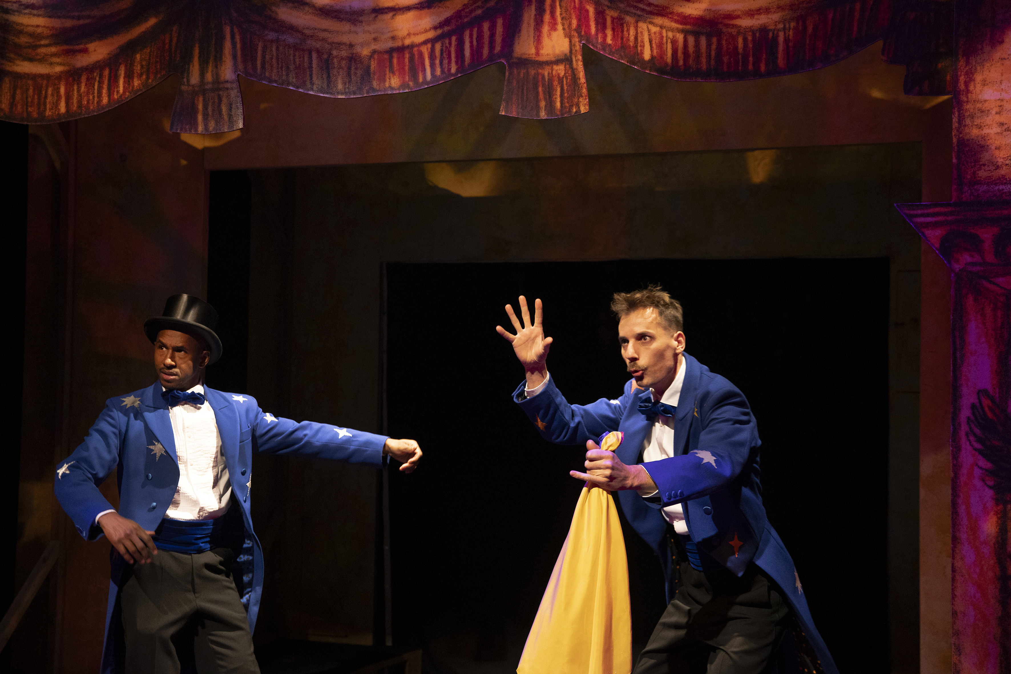 Hier zie je José en Serge op het podium staan. Serge staat links, Serge staat rechts. Ze maken organische bewegingen die je op het goochelpodium zou kunnen verwachten. Ze hebben allebei een blauw rokkostuum aan en een zwarte strik. Serge heeft een geel doek in zijn hand en hij houdt zijn andere hand omhoog.