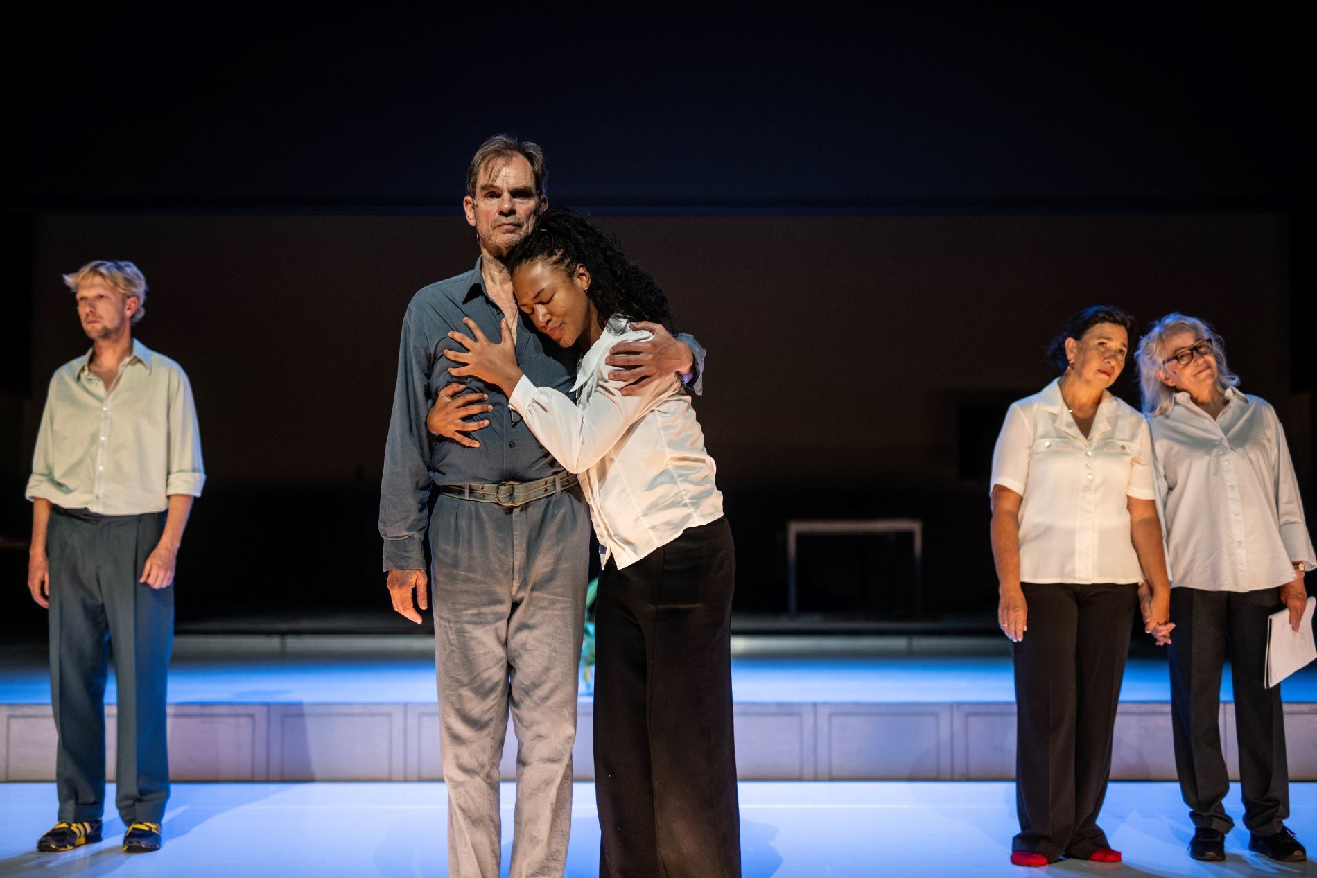 Vijf mensen staan op een podium. Links staat een blonde manier met een overhemd en een blauwe broek voor zich uit te staren. In het midden omhelzen twee mensen elkaar: links een man en rechts een vrouw. De man staart voor zich uit en de vrouw kijkt geëmotioneerd. Rechtsachter staan twee vrouwen die elkaars hand vasthouden. Ze dragen beide een witte blouse en zwarte broek. De linker vrouw kijkt peinzend voor zich uit en de rechter kijkt vrolijk.