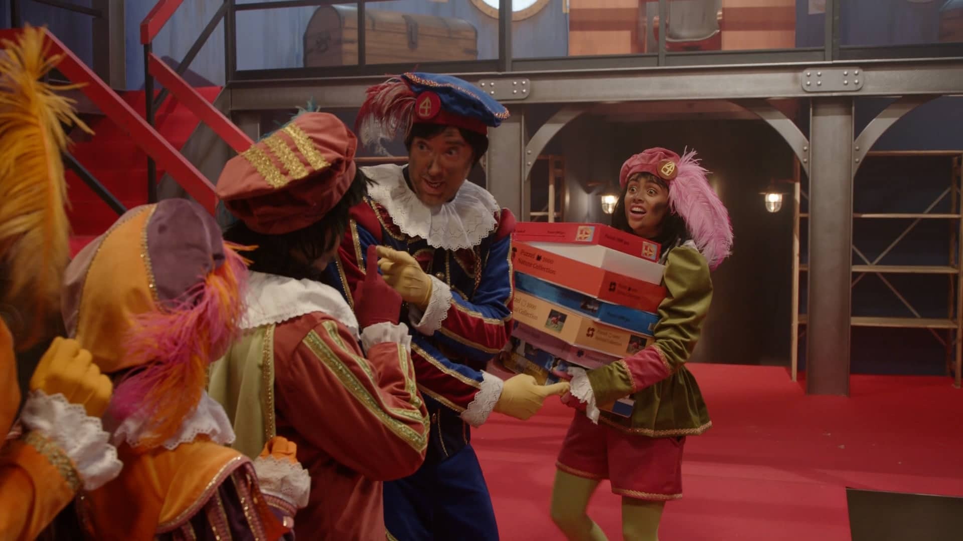 Je ziet 4 mensen in beeld. allemaal in kleurrijke pakjes met mutjes en veren. Een Damen in het kleurrijke pakje, houd puzzeldozen vast. De pakken en personen kunnen gezien worden als pieten van het sinterklaas feest.