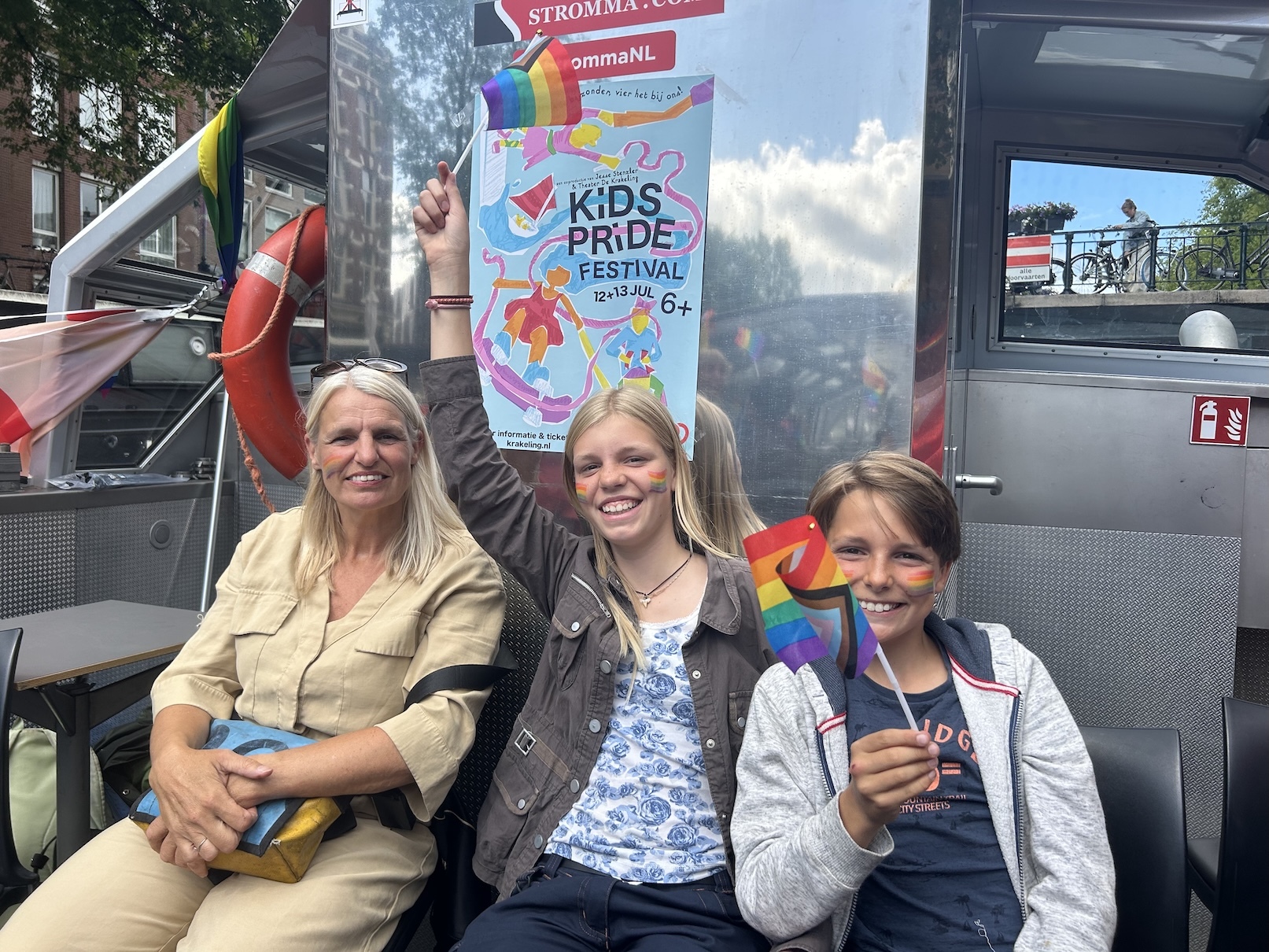 Een moeder en twee kinderen aan boord van de boot. De kinderen zwaaien met regenboogvlaggetjes.