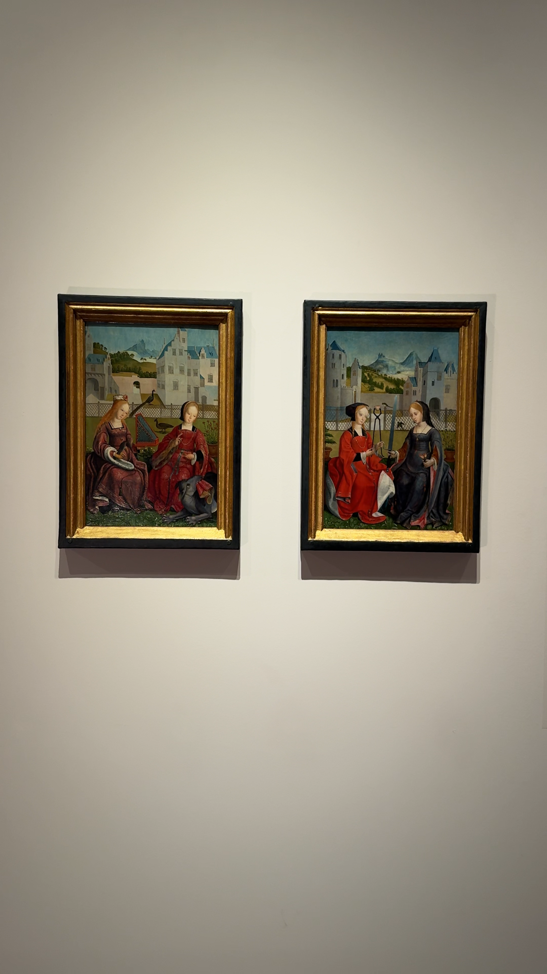 een foto van de twee schilderijtjes die besproken worden in de tekst. Het zijn twee kleine schilderijtjes met vrouwelijke heiligen erop. Ze zitten in gouden lijstjes en hangen naast elkaar. De vrouwen zitten in een soort kloostertuin en hebben allemaal mooie jurken aan met veel goud.