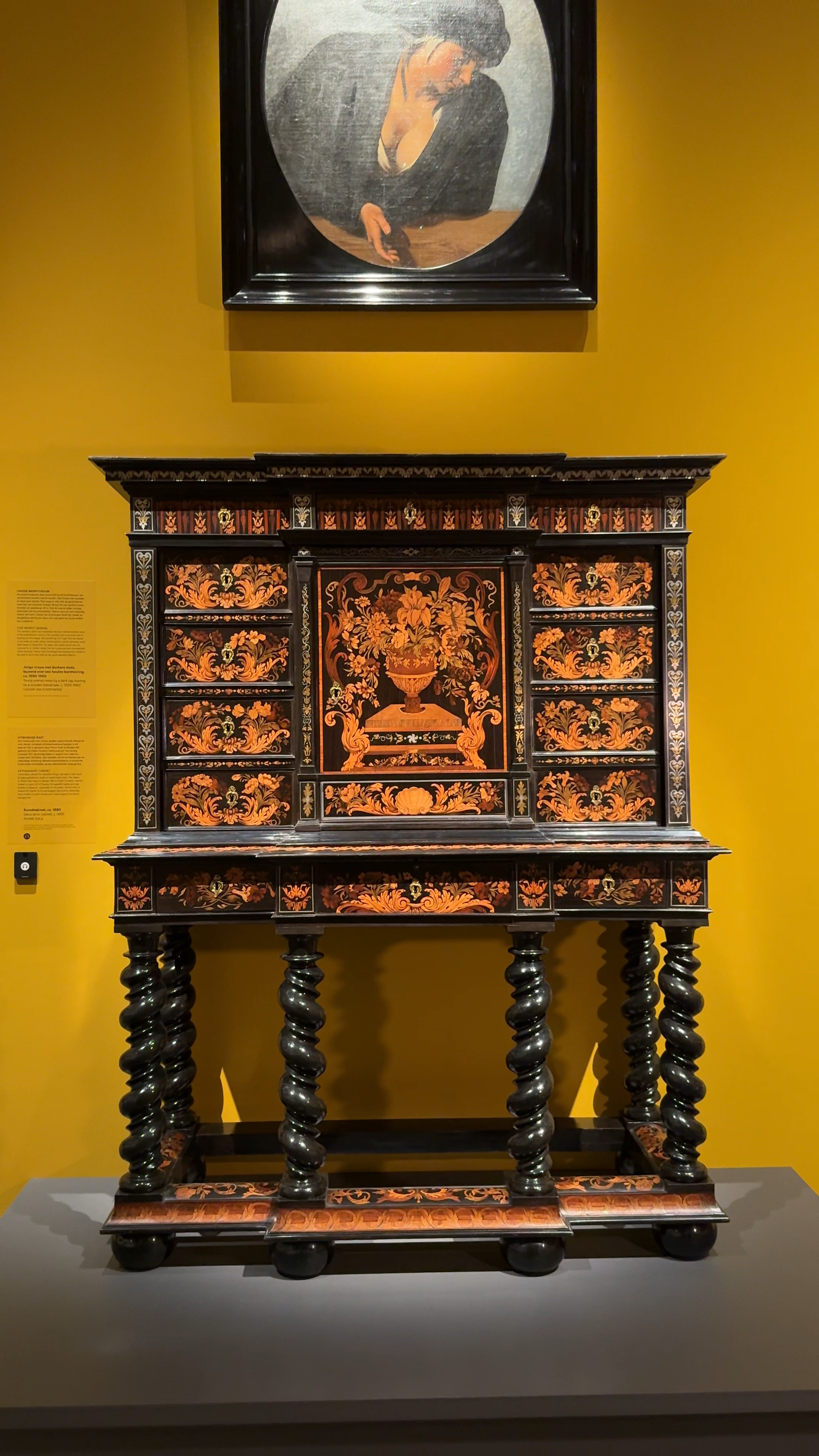 Een foto van de kast die wordt besproken. Het is een ebbenhouten kast, die op poten van ongeveer een meter staat. Er is een bloemenpatroon van hout ingelegd. Hij staat voor een gele muur en erboven hangt een schilderij.