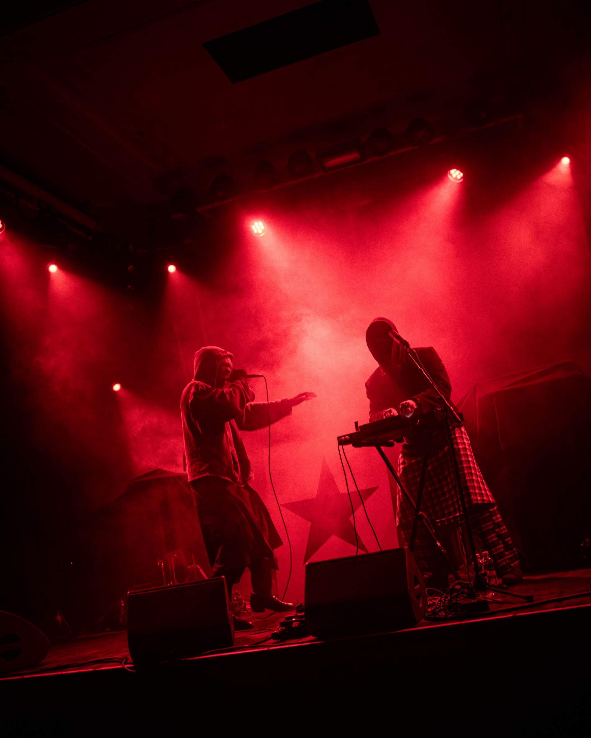 Artiest Isaï staat op het podium op te treden, naast hem staat een man. Het decor bestaat uit rode lampen, rook en een zwarte ster op de achtergrond.