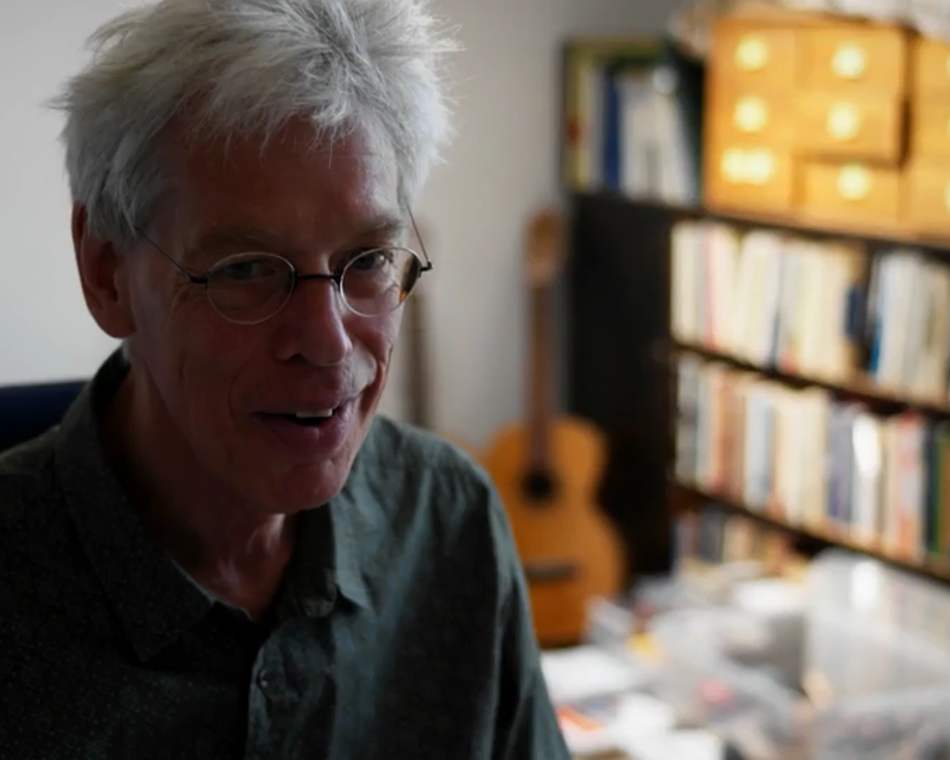 Joël Bons zit in zijn kamer, met achter hem in de hoek een elektrische gitaar en daarnaast een boekenkast die vol staat. Hij heeft een donkergroen overhemd en een ronde bril op.