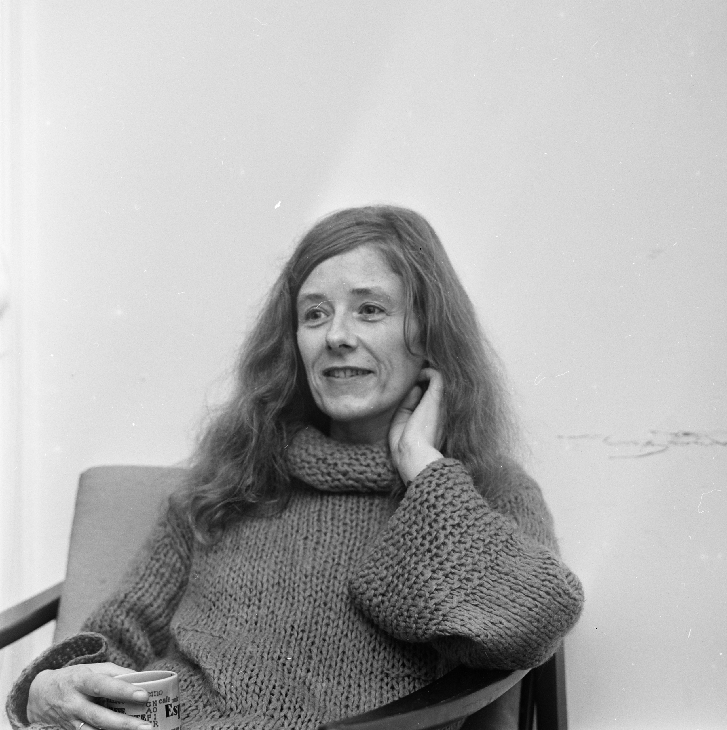Zwartwitfoto van Joke Smit die op een stoel zit met een mok in haar hand. Ze lacht, heeft haar hand in haar nek en draagt een gebreide coltrui.