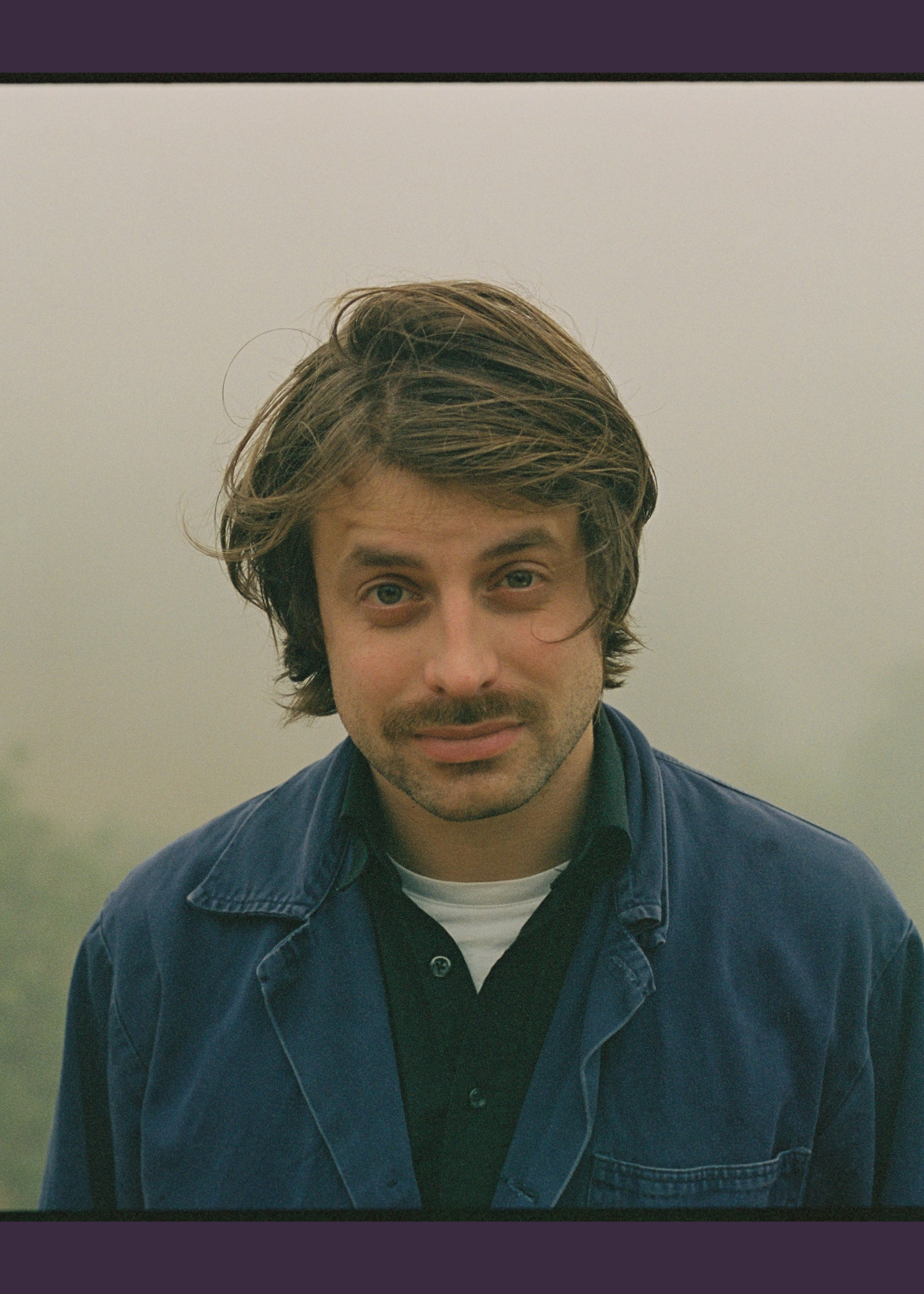 Een portretfoto van Joost Oomen. Een jongeman met wilderig haar, buiten gefotografeerd. Hij heeft een snor en draagt een blauwe jas.