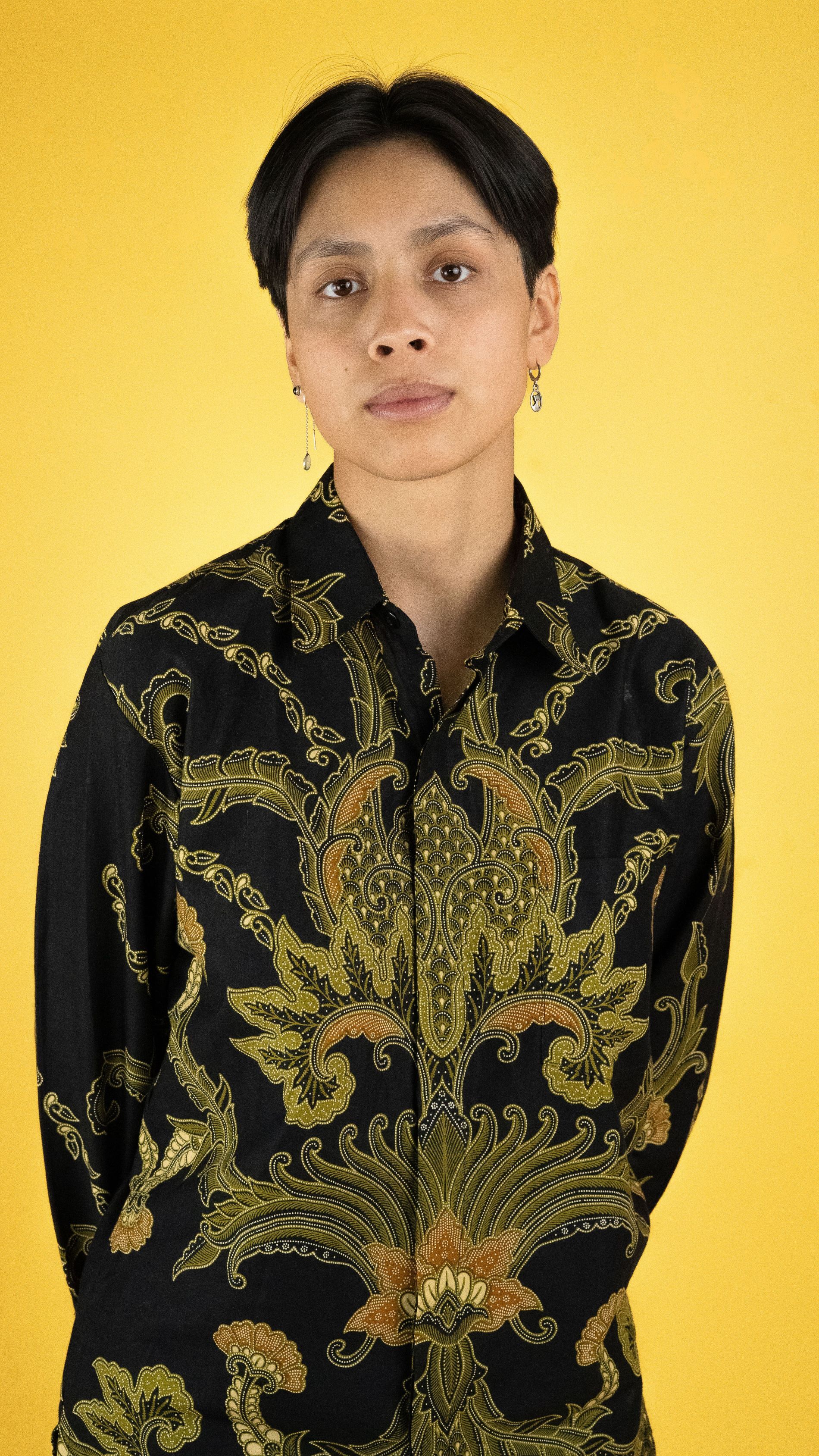 Vrouw met kort zwart haar staat voor een gele muur. Ze kijkt in de camera en draagt een batikhemd met groene bloemen.