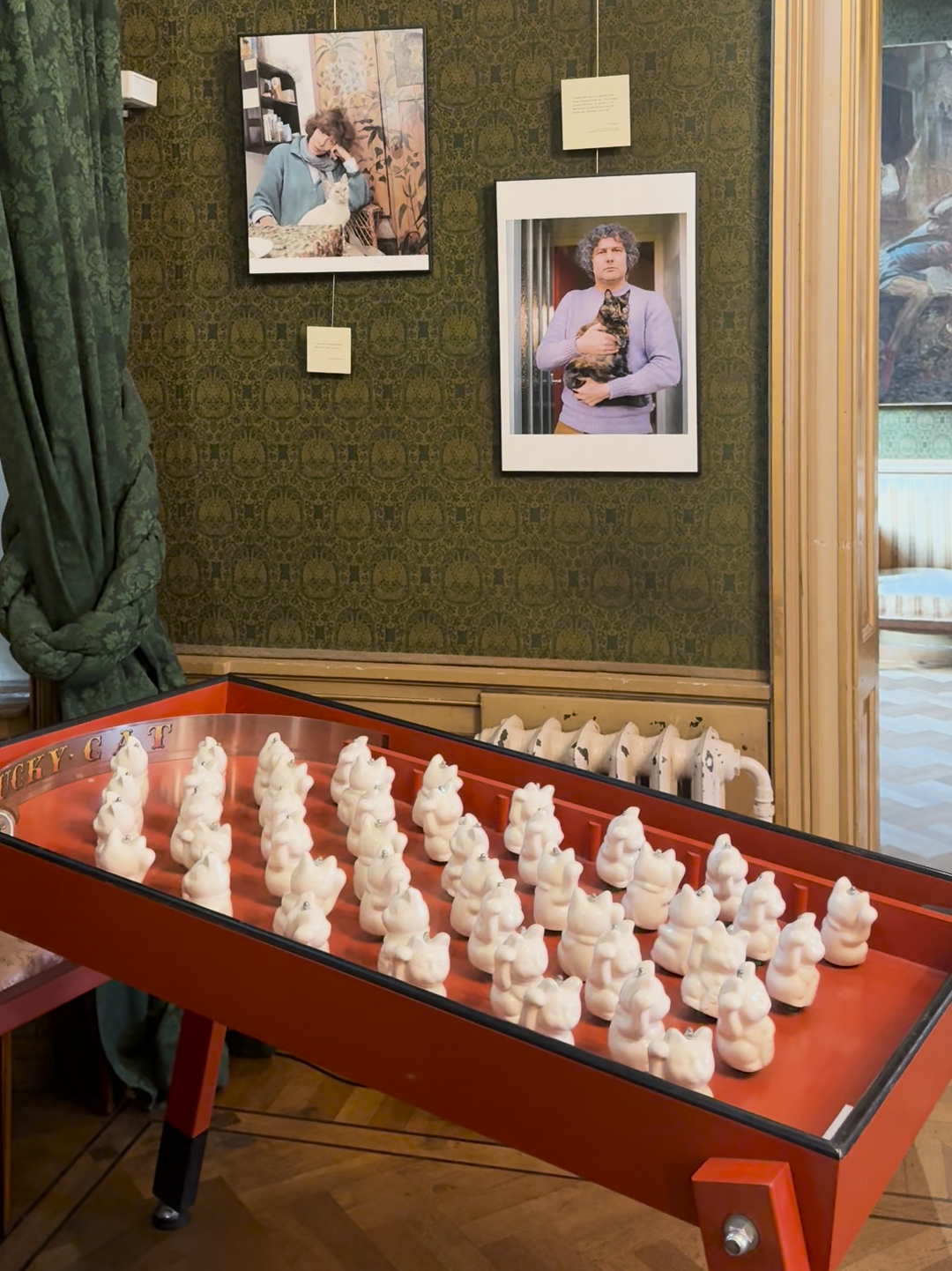 Vooraan staat een rode speltafel met heel veel Chinese gelukskatjes. Achter zie je een groene muur met daaraan portretfoto's van de tentoonstelling over kunstenaars en hun katten in de fotografie.