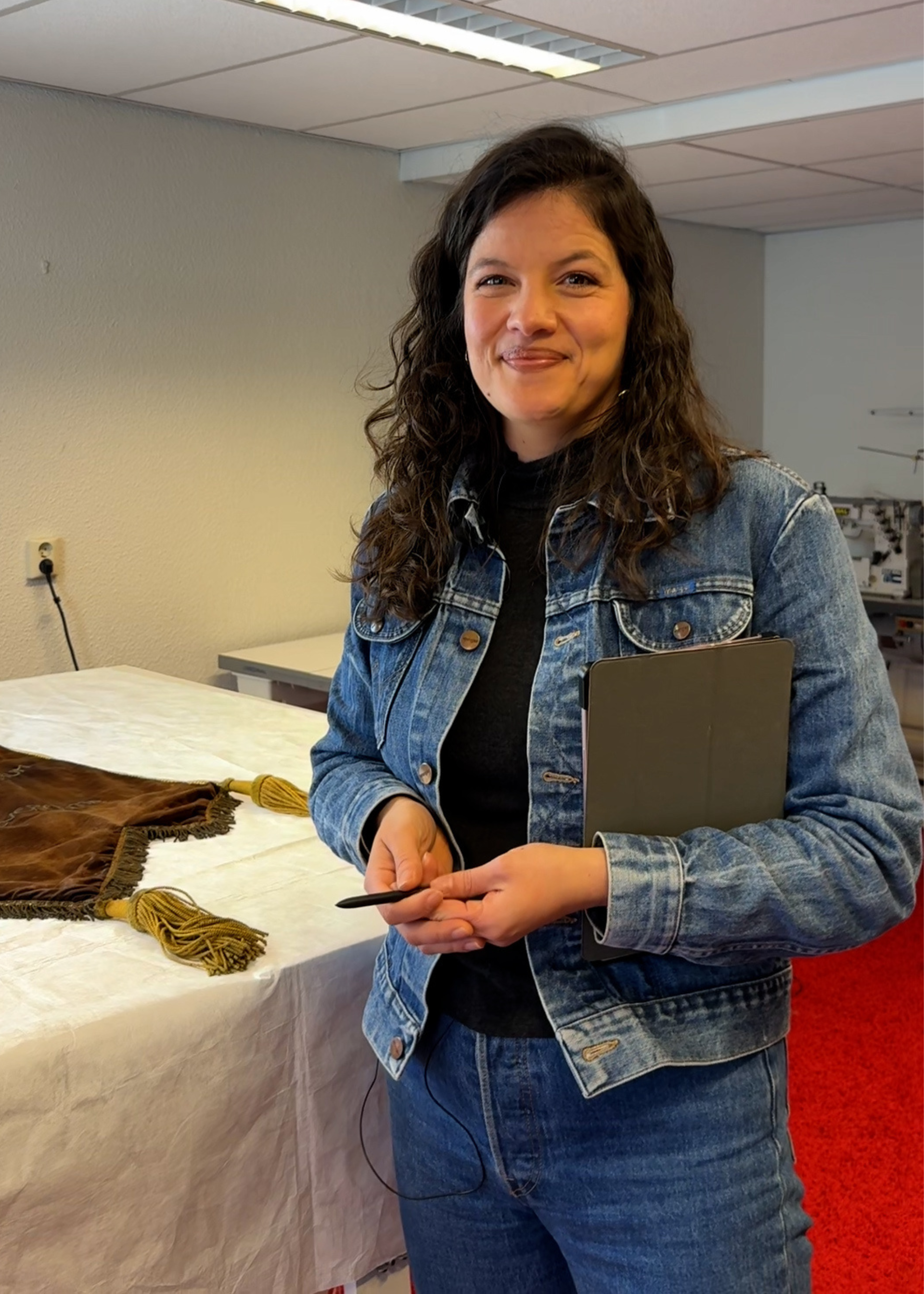Lorena in haar atelier. Ze heeft een spijkerpak aan en los bruin haar. Ze lacht naar de camera. Ze heeft een tablet en pen in haar hand.