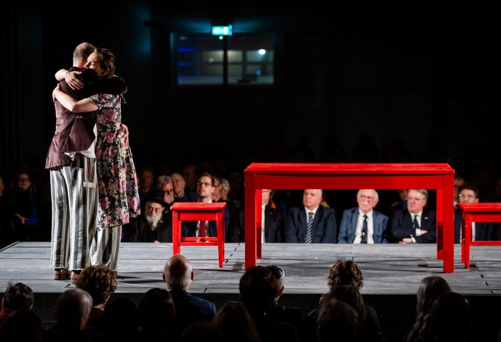 Op de foto zijn twee knuffelende personen op een podium te zien. Een persoon, de vrouw, heeft een bloemetjesjurk aan tot over haar knieën. Daaronder draait ze een loszittende grijs-gestreepte broek. De man draagt een bruin colbert. Ook hij draait dezelfde soort grijs-gestreepte broek. Naast hen op het podium staan een rode tafel en stoel.