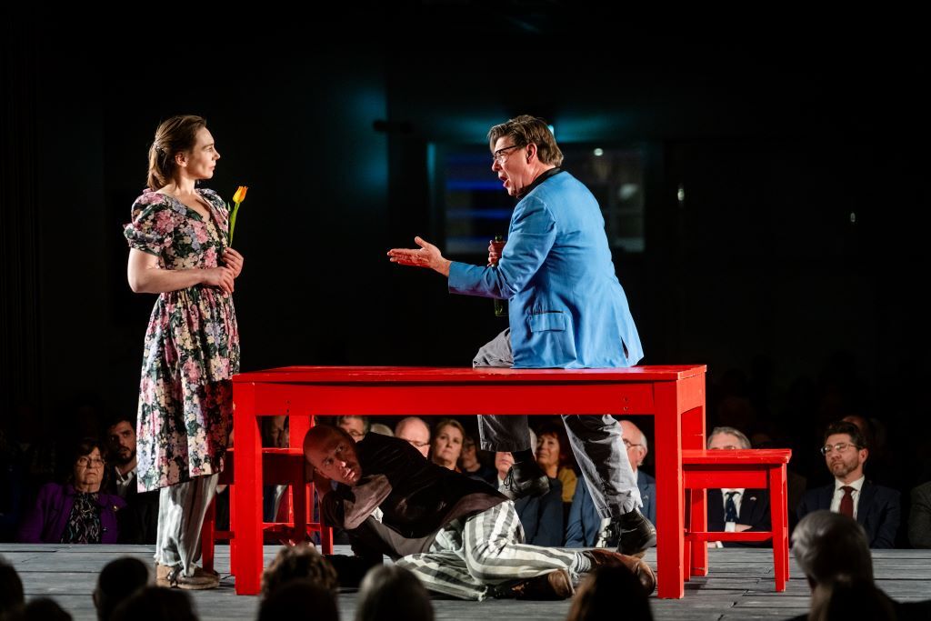Hier zie je op een podium drie mensen staan rondom, op of onder een rode tafel en twee rode stoelen. Links van de tafel staat een vrouw met een bloemetjesjurk aan en daaronder een gestreepte broek. Ze heeft een bloem in haar handen. Op de tafel zit een man met een blauw colbert en daaronder een gestreepte broek. Hij heeft zijn mond open want hij zingt en hij wijst met een hand naar de vrouw. Onder te tafel ligt een man op zijn zij. Hij draagt een bruin colbert en dezelfde gestreepte broek.