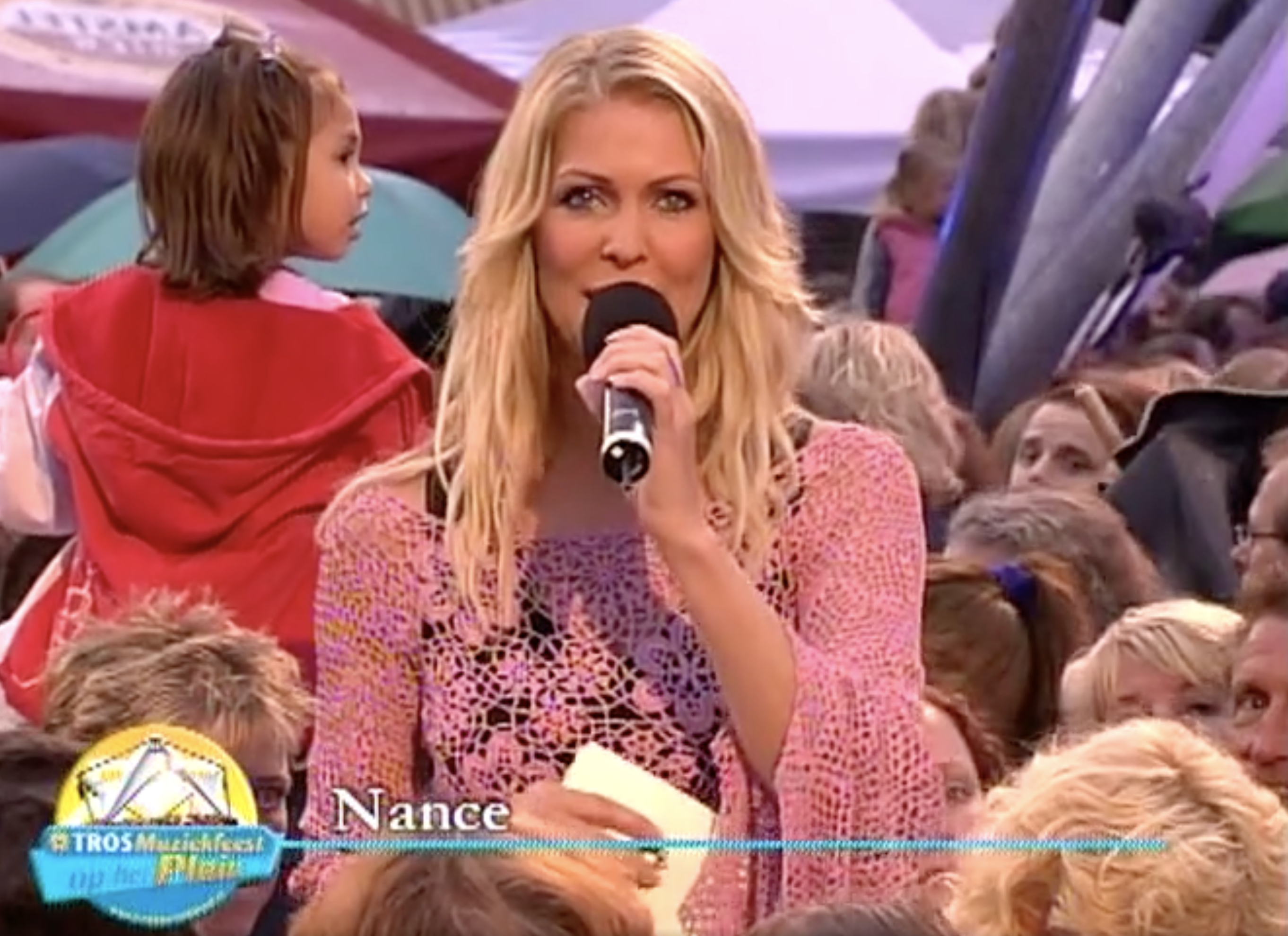 Presentatrice Nance. Ze praat in een microfoon en heeft een roze top aan. Onder in beeld zien we een naambalkje staan met Nance en het tros muziekfeest logo. Ze staat middenin het publiek. Het is een beeld uit de uitzending van 2005 en dat is ook te zien aan het logo en de beeldkwaliteit.