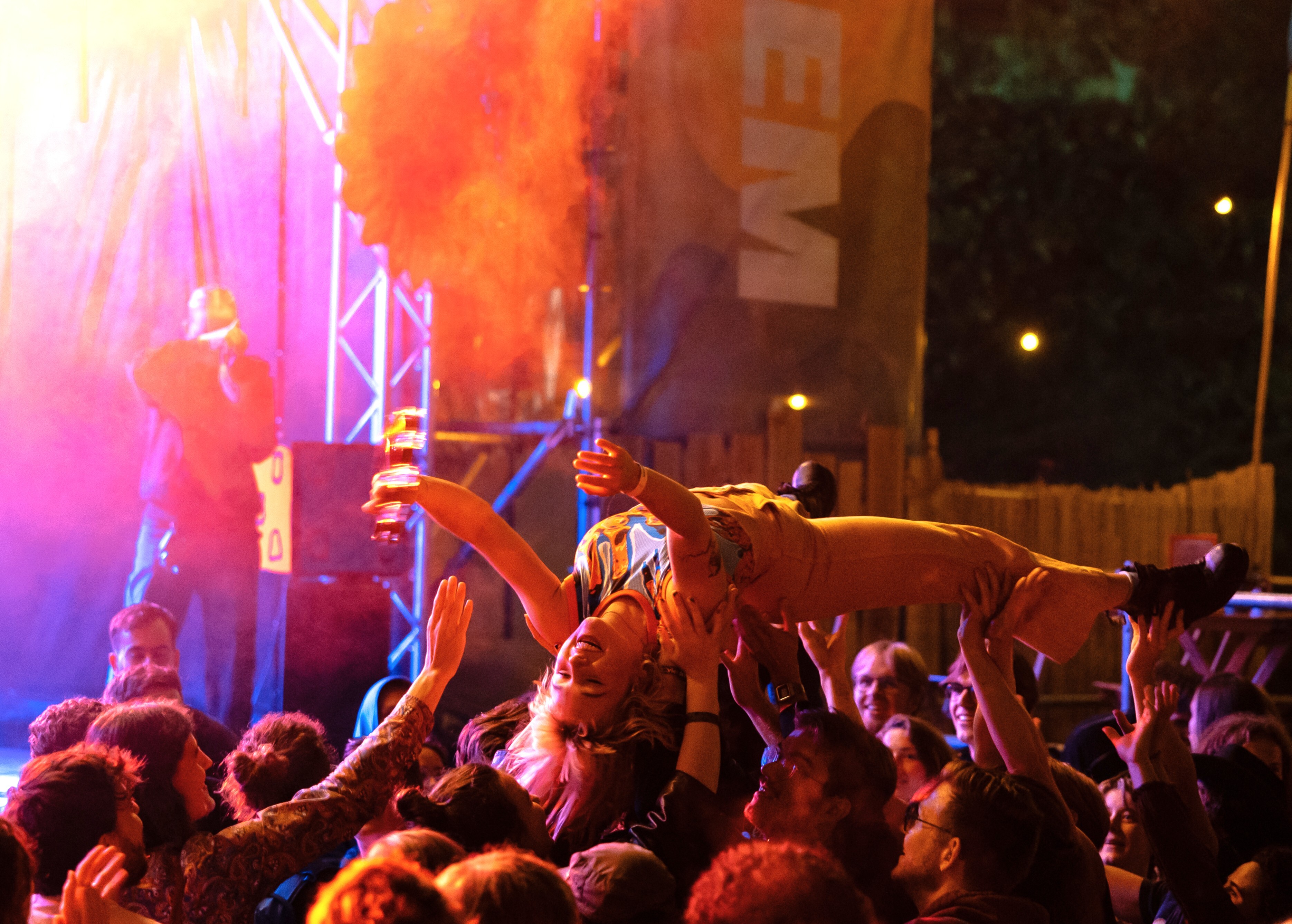 Nelleke crowdsurft over het publiek, ze heeft haar handen omhoog en wordt gedragen door tientallen mensen. De lichten op het podium schijnen deels in de camera waardoor er in de linkerhoek een geel lichteffect ontstaat.