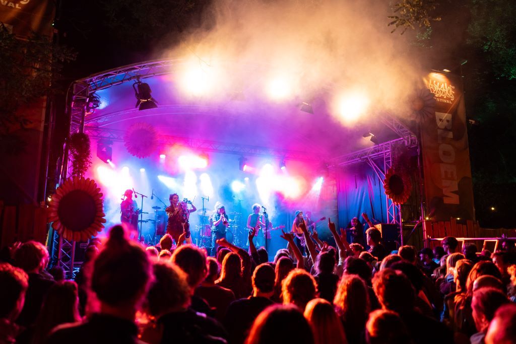 De band PARRA.DICE staat op een podium. Er worden felle lichten op het publiek van ongeveer 50 mensen geschenen. Je ziet alleen hun achterhoofden.