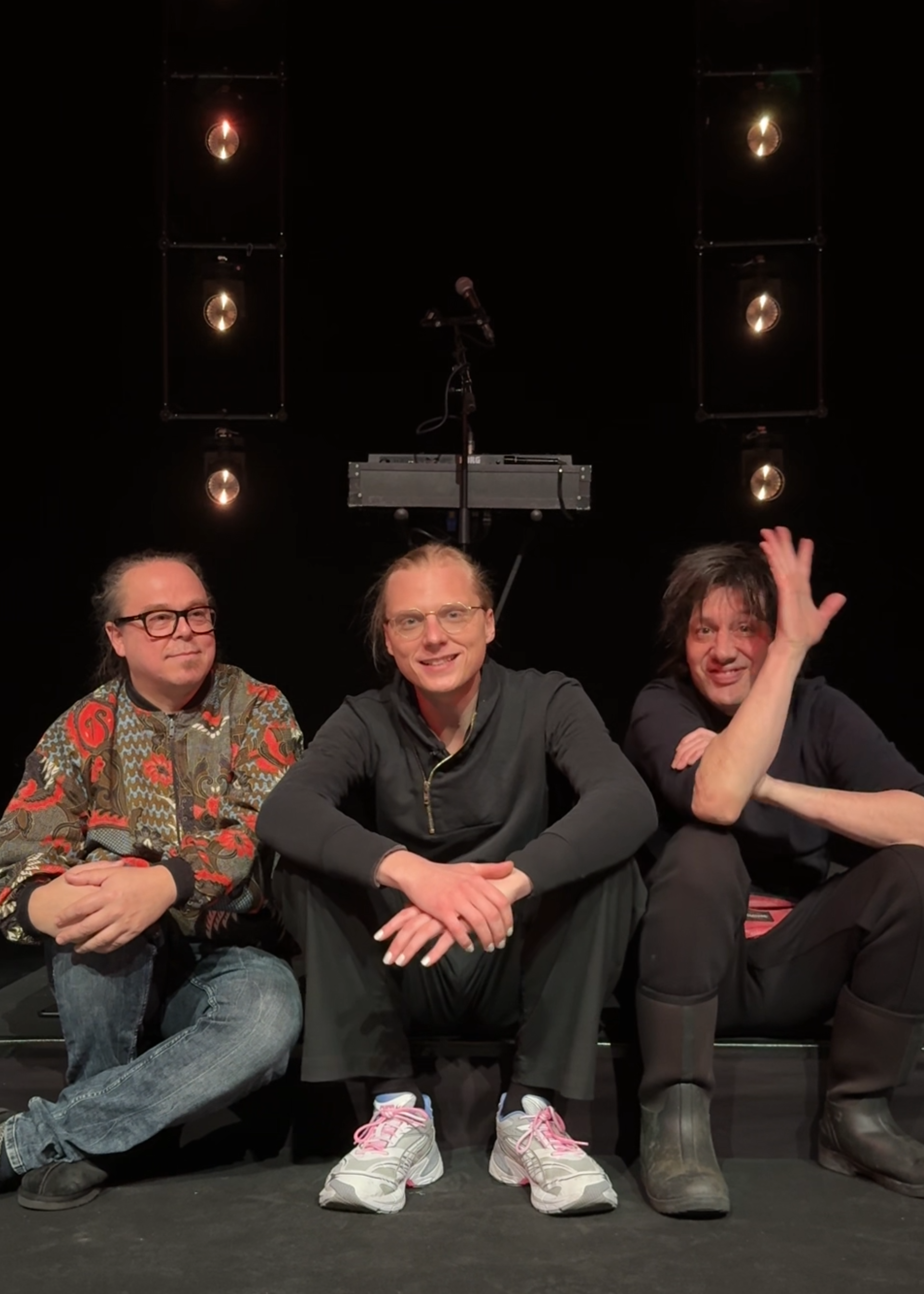 De drie leden van de band Prins S en de Geit zitten op een klein podiumpje en kijken naar de camera. Ze lachen allemaal vriendelijk en de meest rechter zwaait.