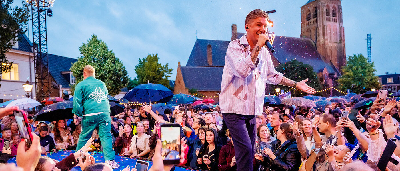 Zanger Mart Hoogkamer op het podium in Moegestel. Op de achtergrond zie je een kerk en heel veel publiek, met paraplu's.