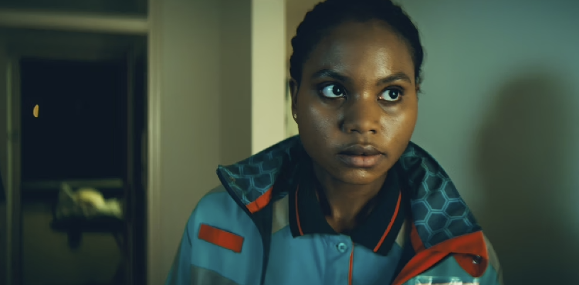 Alima staat in een huis met een ambulance jas aan en kijkt naar iets buiten beeld