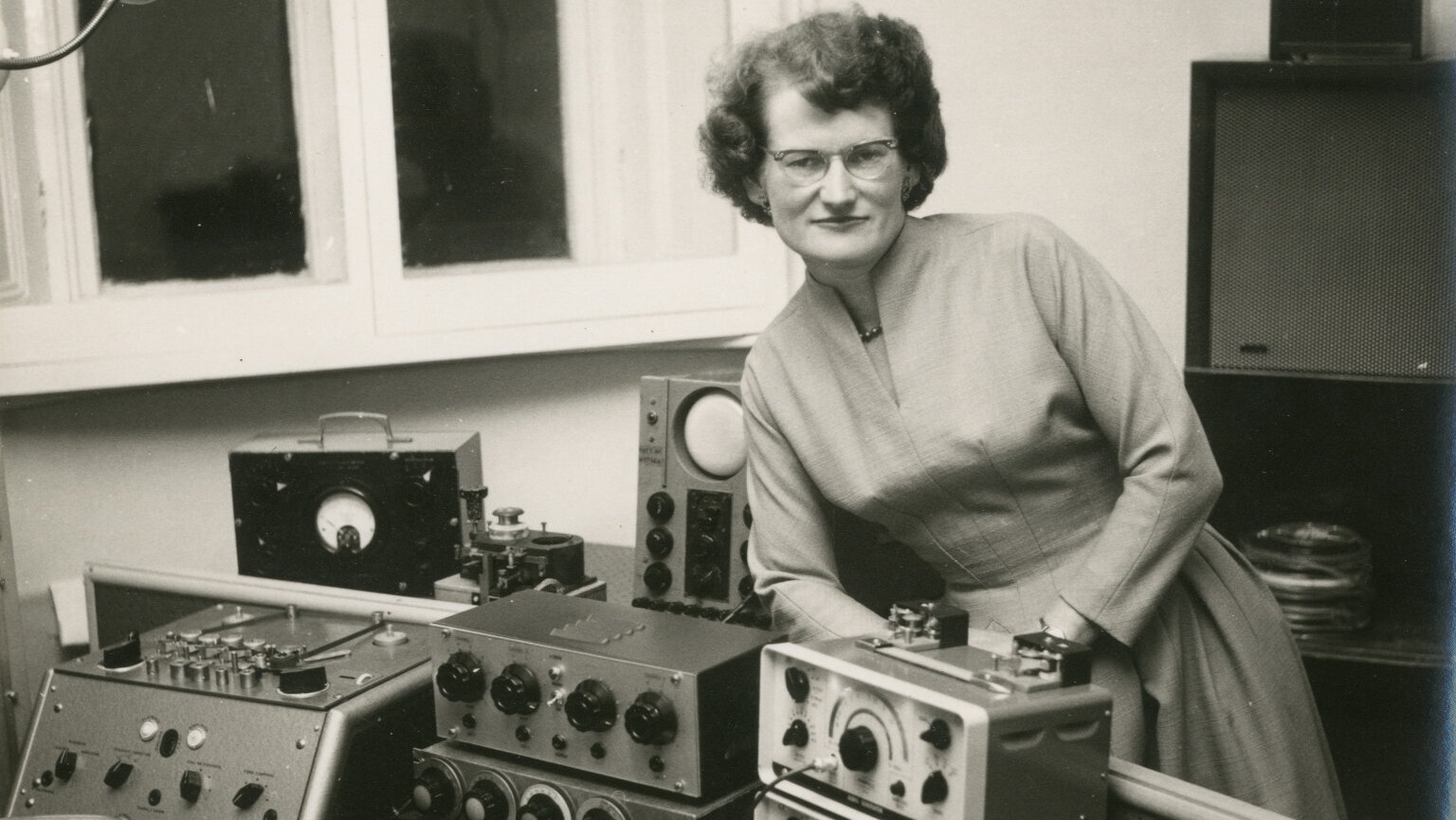 Componist op zwart wit foto omringd door apparatuur, waarop ze elektronische muziek componeert.