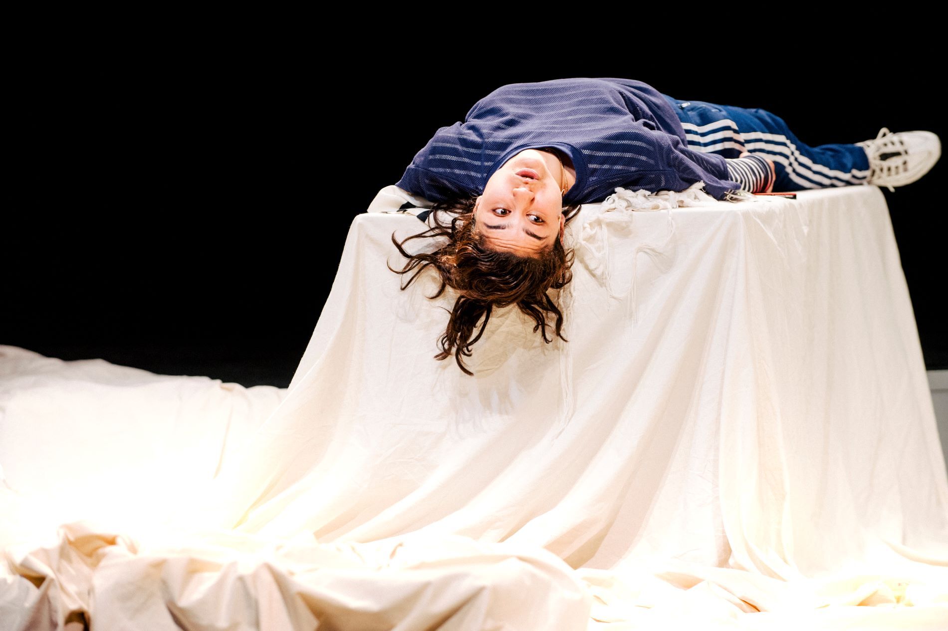 Actrice Soumaya ligt in blauwe kleding met haar rug op een tafel met een groot wit doek. Haar hoofd hangt over de tafel heen en ze kijkt naar rechts.