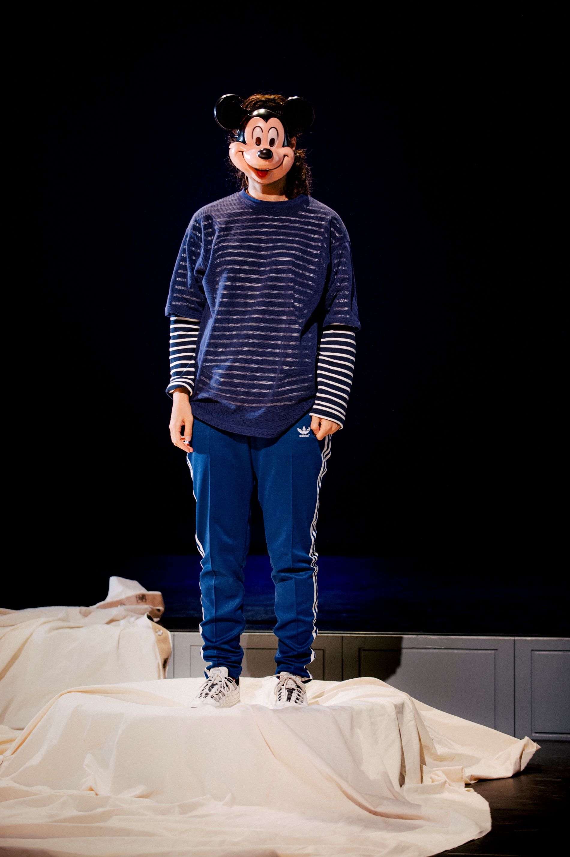 Actrice Soumaya staat op een trampoline die bedekt is door een wit kleed. Ze draagt een blauw gestreept shirt, blauwe broek en een Mickey Mouse-gezichtsmasker.