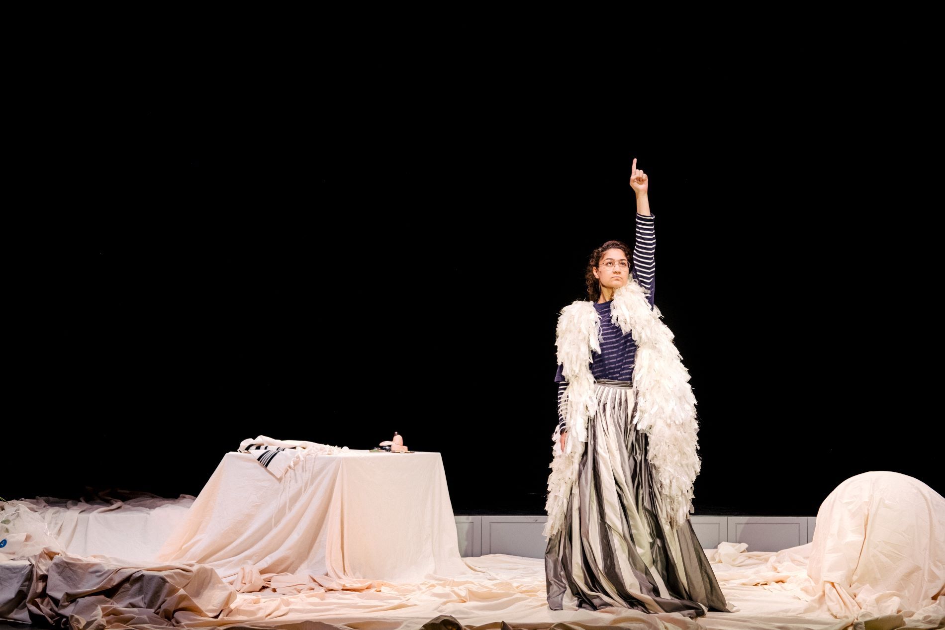 Actrice Soumaya staat op het podium met haar vinger in de lucht. Ze draagt een gestreept shirt, een witte boa en een lange grijze rok. De vloer is bedekt met witte kleden.