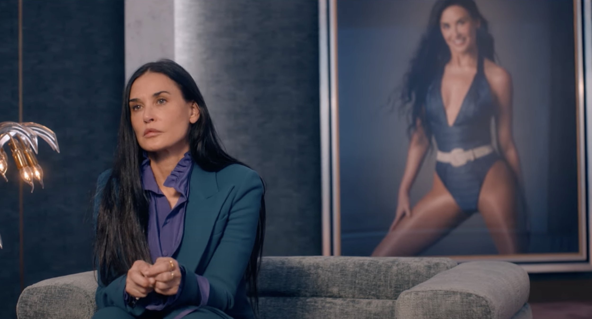 Een stil uit de film The Substance. Een treurig kijkende vrouw zit op een bank. ze heeft lang zwart haar en achter haar hangt een geposeerde studio-foto van haar in badpak.