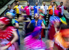 Zwarte vrouw in felblauwe jurk met een hoofddoek staat met haar handen tegen elkaar aan. Om haar heen dansen mensen in roze, oranje en blauwe kleding.