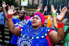 Zwarte vrouw in felblauwe jurk met een roze hoofddoek en rode armbanden staat met haar handen omhoog. Achter haar staan mannen in in roze, oranje en blauwe kleding die dezelfde pose doen.