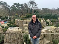 Zandkunstenaar Maxim Gazendam staat voor een railing. Achter hem zie je verschillende zandkunstwerken uit de tentoonstelling in Garderen, waaronder een groot mensenhoofd.