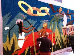 Er wordt door twee vrouwen gewerkt aan het wandtapijt. Op het tapijt staan twee personen, gebroken ketens, nootmuskaat en een palmboom.